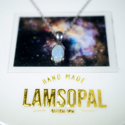 A silver chain necklace with an oval light blue opal pendant displayed on a branded card. The card features a cosmic galaxy background and gold foil text reading 'HAND MADE LAMSOPAL NATURAL SPA'.
