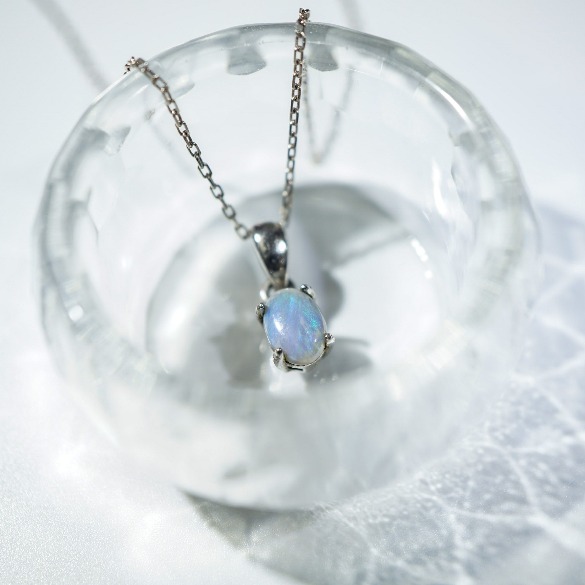 A silver chain necklace with an oval iridescent opal pendant, displayed inside a clear glass bowl on a white textured surface.