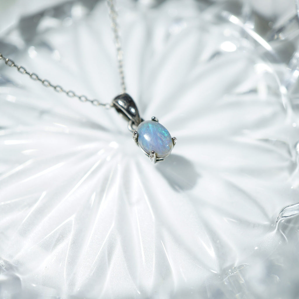 A silver pendant necklace featuring an oval iridescent opal gemstone, resting on a textured white glass surface.