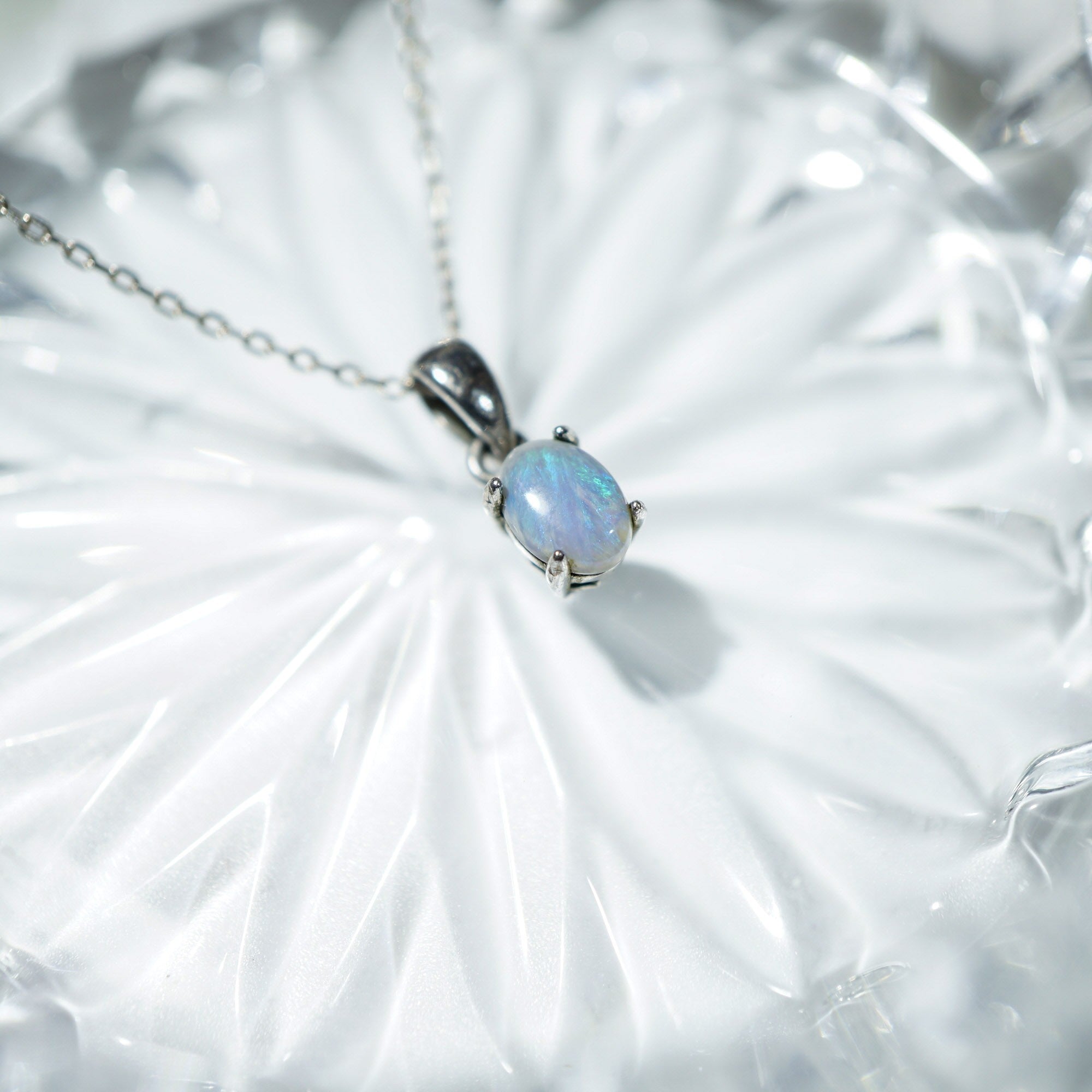 A silver pendant necklace featuring an oval iridescent opal gemstone, resting on a textured white glass surface.