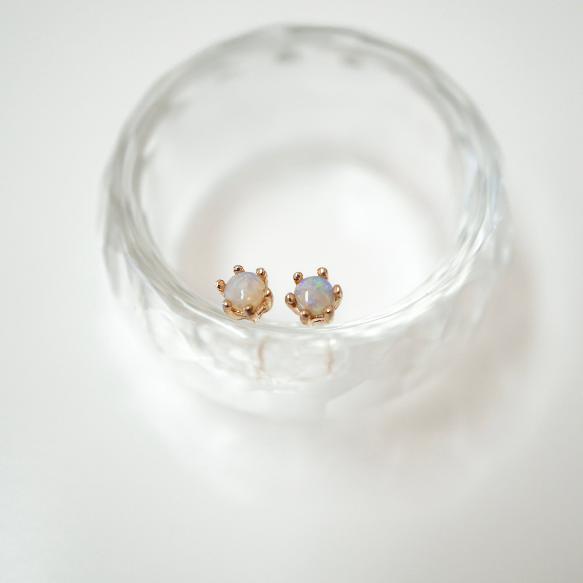 A pair of small gold stud earrings with round iridescent white stones, displayed inside a clear textured glass bowl on a white background.