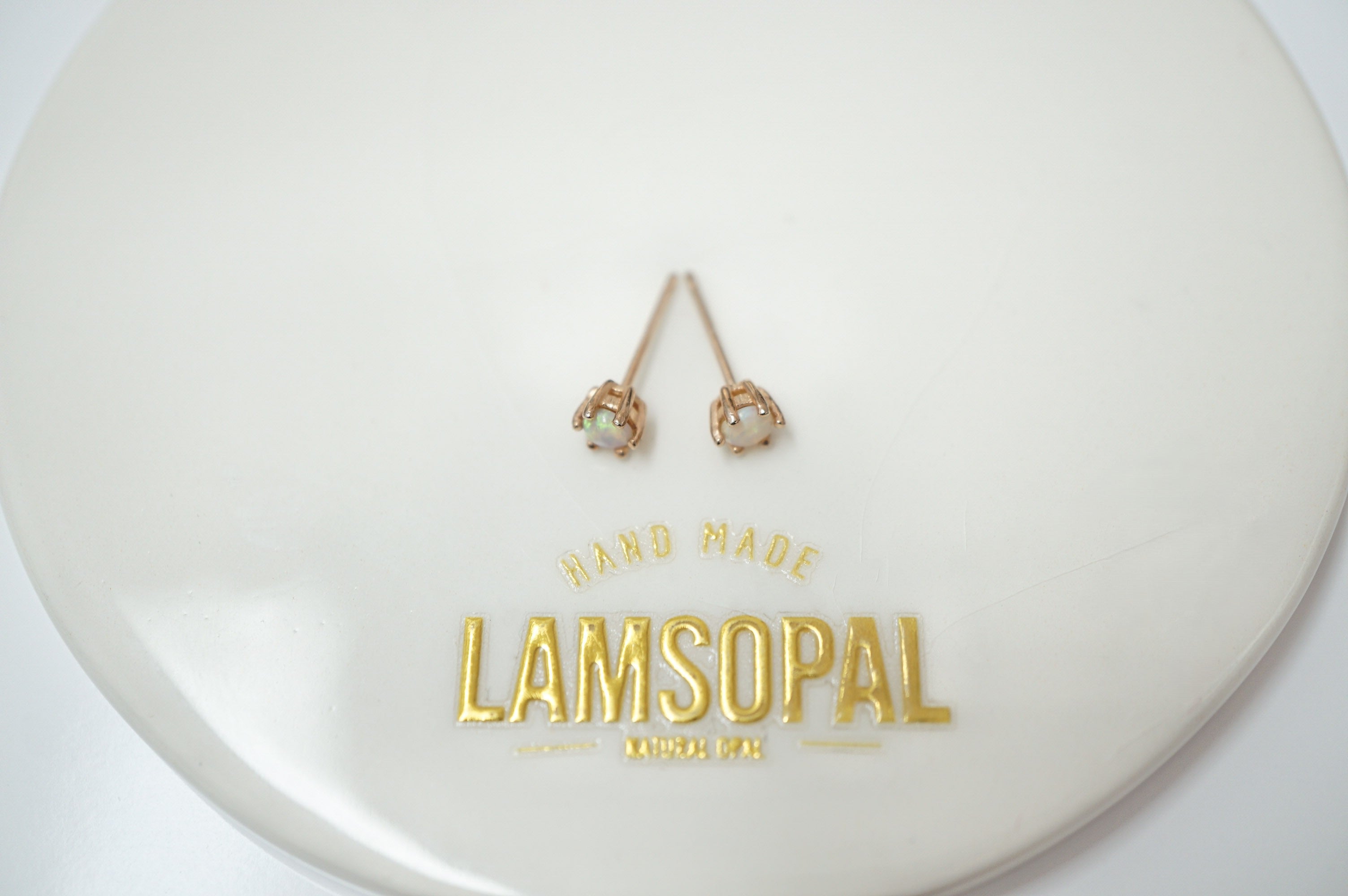 A pair of small gold stud earrings with oval light green stones, displayed on a glossy white circular stand with gold 'LAMSOPAL HAND MADE' branding.