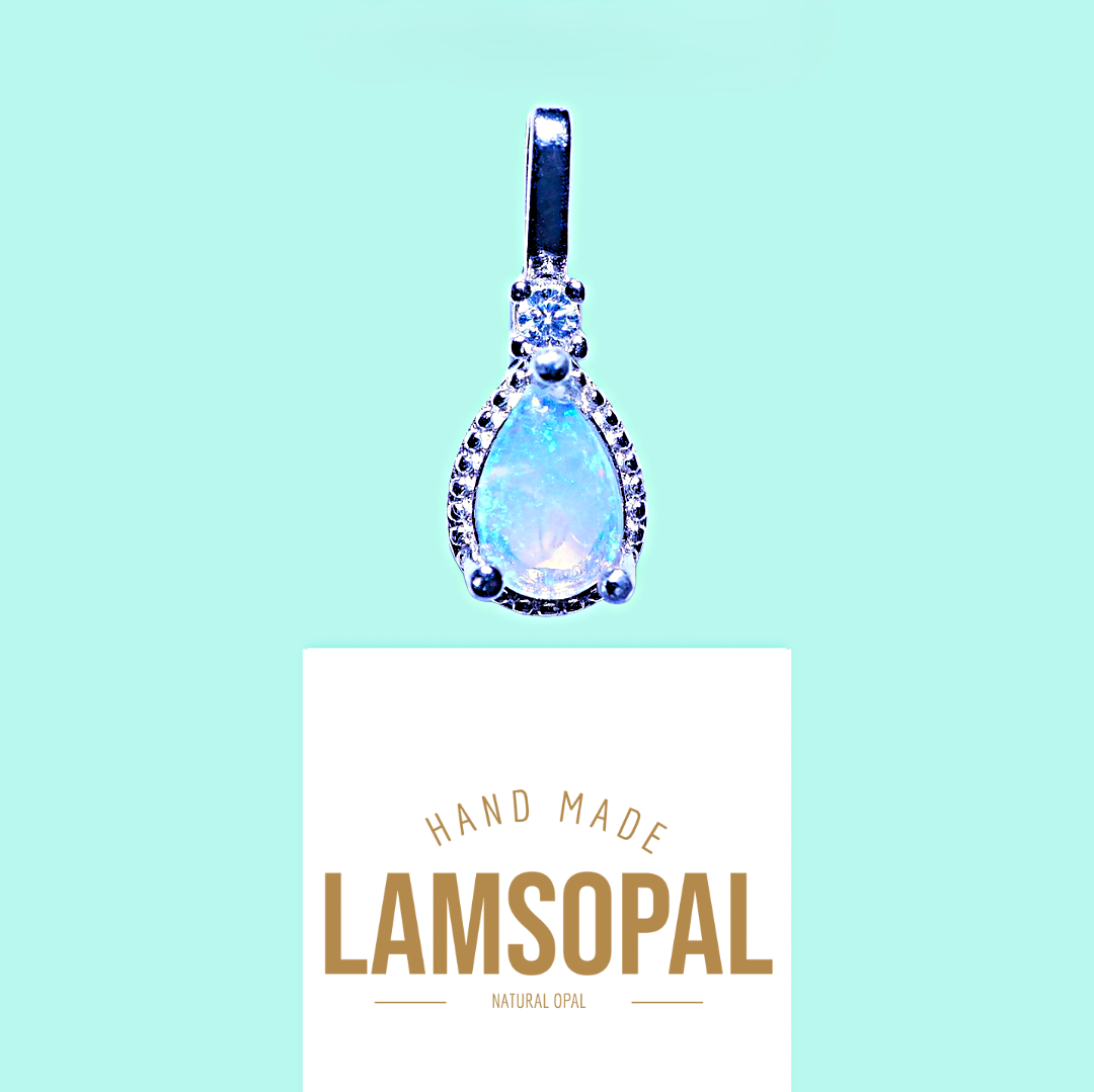 A teardrop-shaped Lamsopal pendant with a light blue iridescent center and silver-toned bezel set with small clear stones, shown above a white card printed with the brand name LAMSOPAL on a light aqua background.