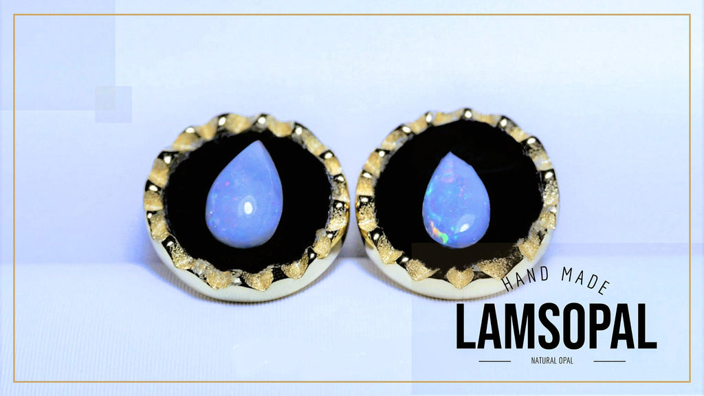 A pair of Lamsopal natural opal stud earrings with teardrop-shaped light blue opal centers set in a gold bezel with a black background, shown on a light blue background with a gold border.