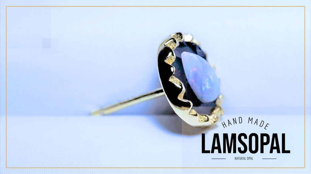 A handmade Lamsopal ring featuring a natural opal gemstone with iridescent blue and purple hues, set in a gold-toned bezel with black accents, on a light blue background with a gold border.