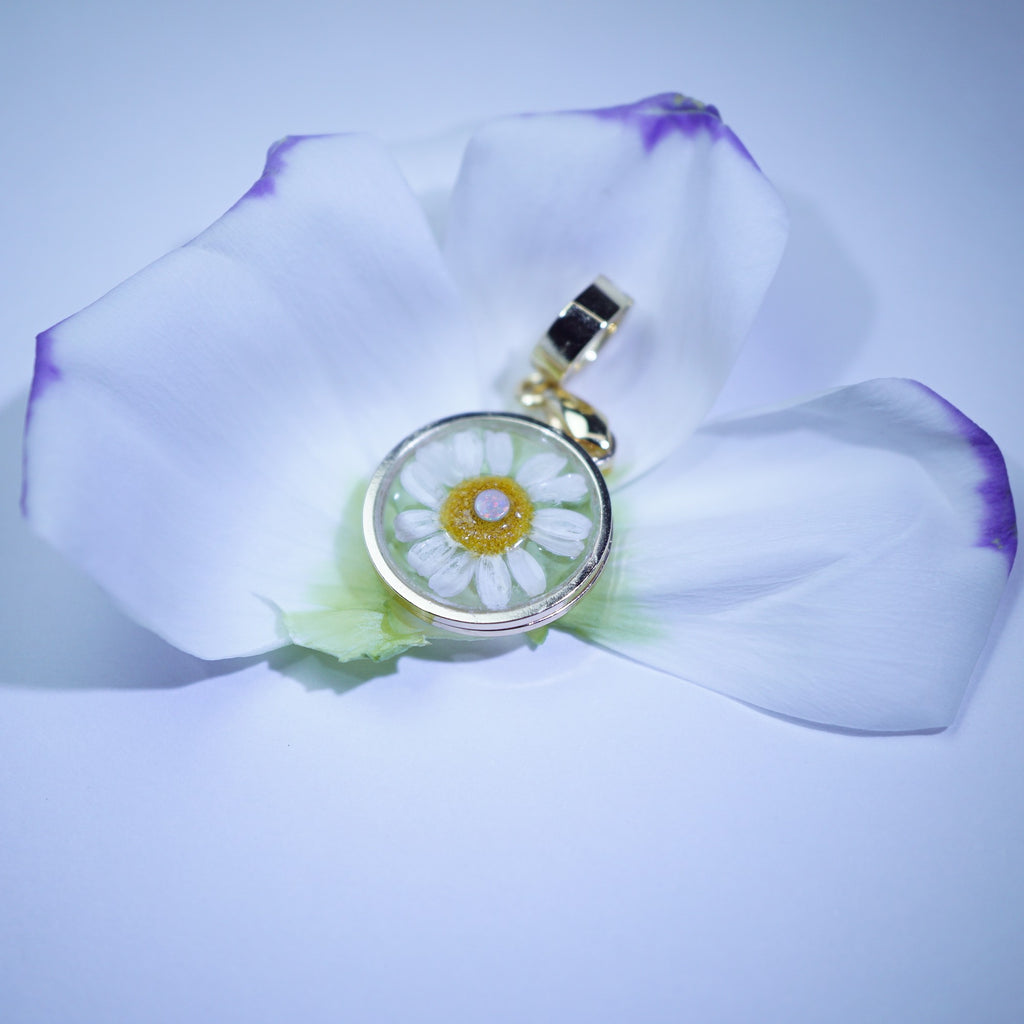 A round pendant charm featuring a white daisy flower with a yellow center, set in a gold-toned bezel and attached to a gold bail, resting on a white flower petal.