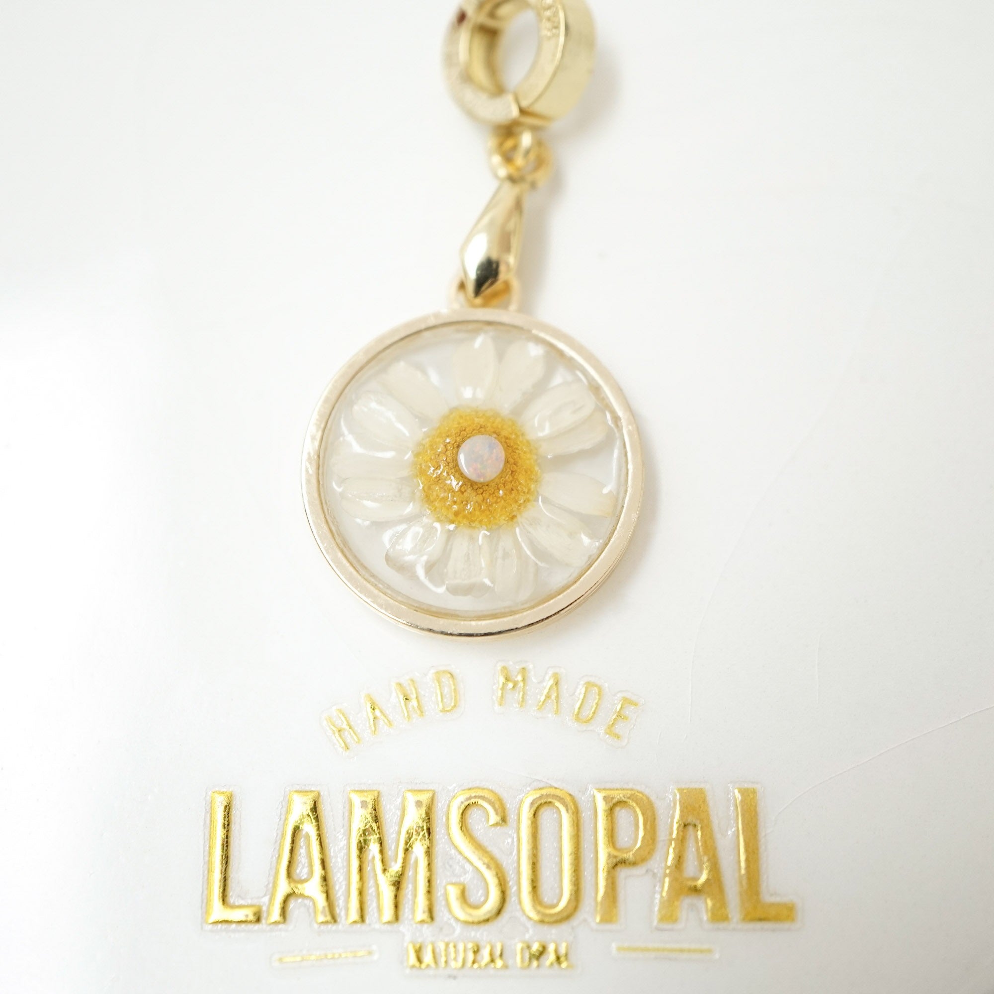 A gold-tone circular pendant charm featuring a white daisy flower with a textured yellow center and a small white pearl, shown on a white background with the brand name 'LAMSOPAL' below.