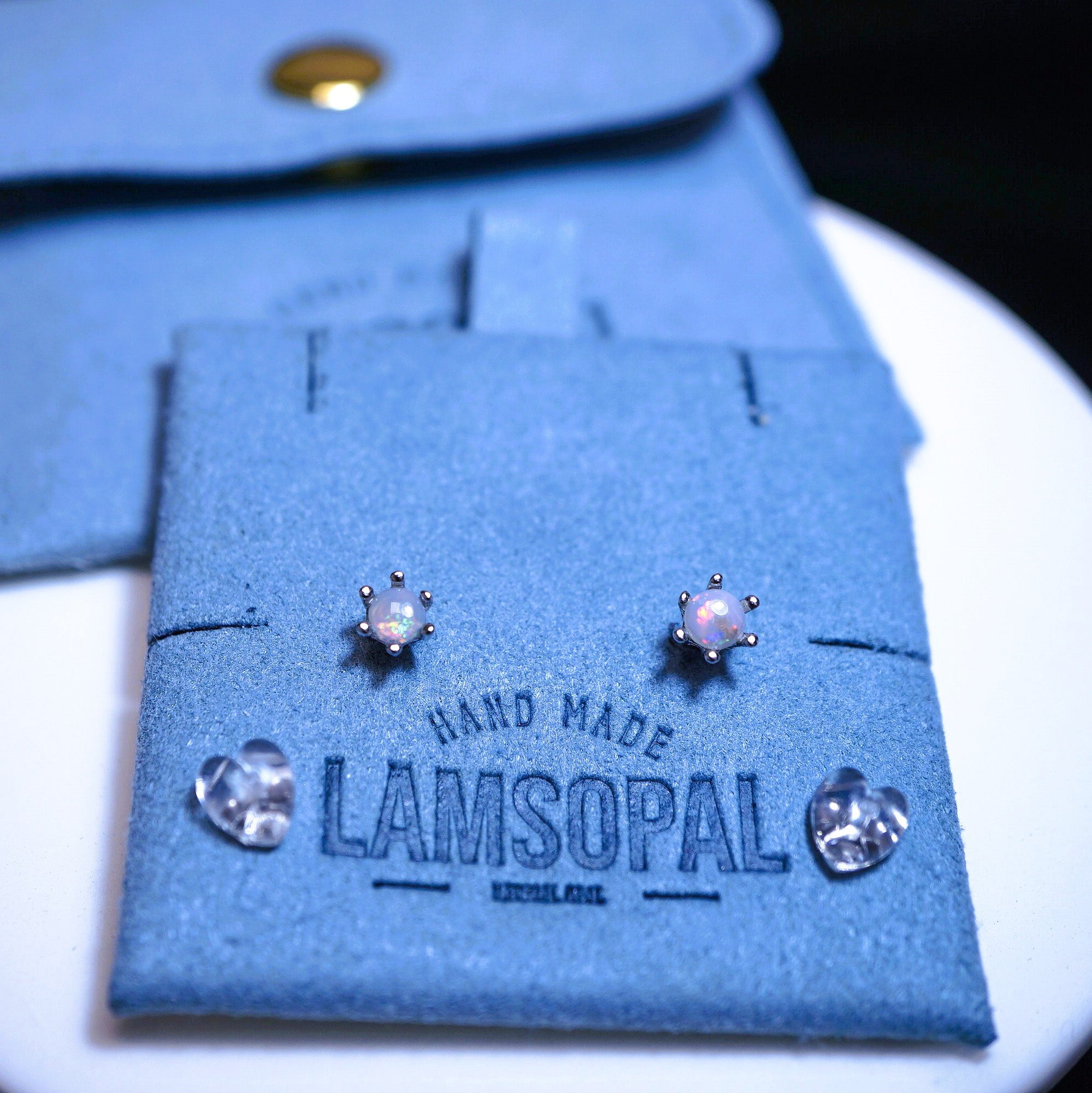 A pair of small iridescent opal stud earrings displayed on a blue velvet jewelry card with the brand name Lamsopal, alongside two clear crystal-like accent stones, with a matching blue pouch in the background.
