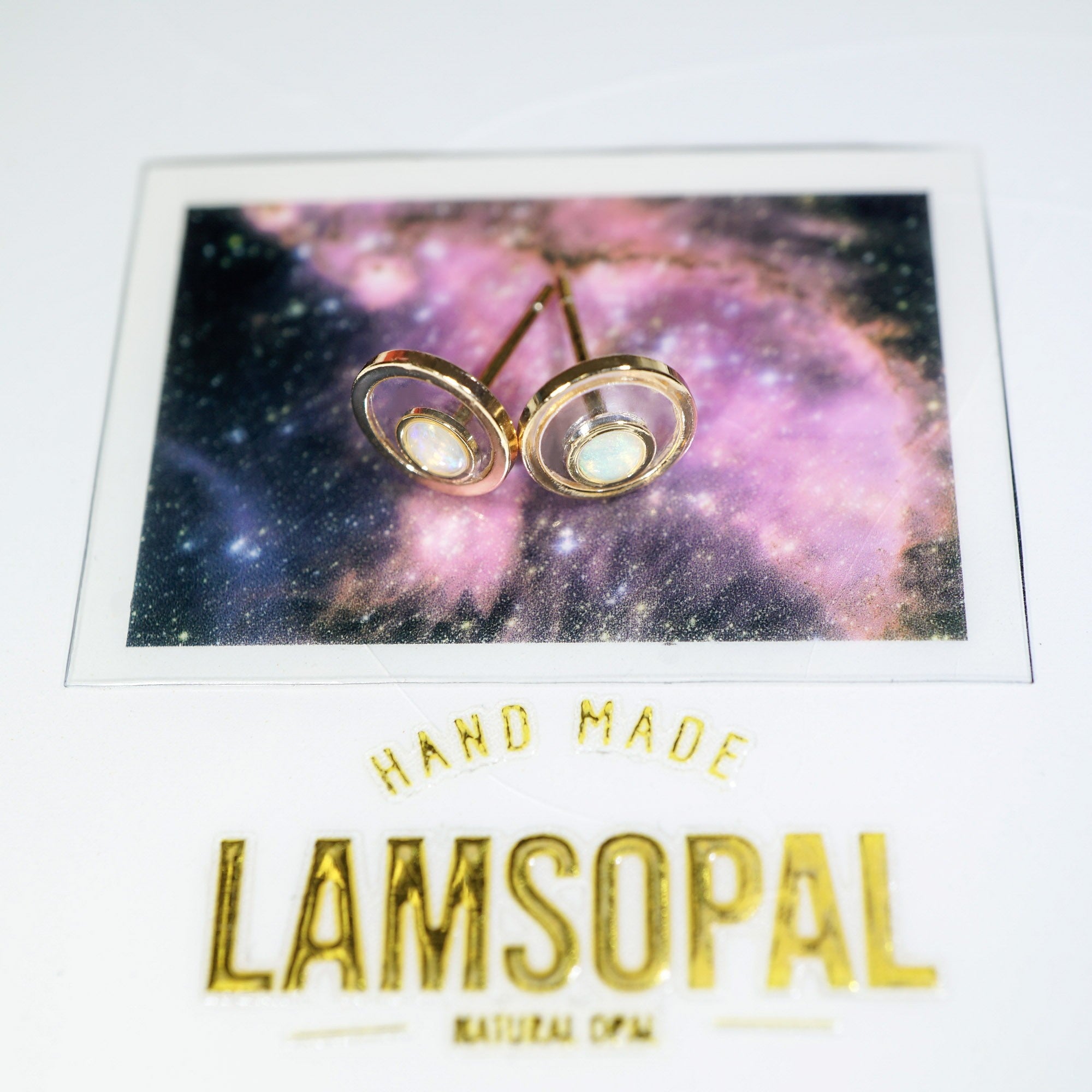 A pair of gold-toned double-circle stud earrings with white inlay, displayed on a printed card featuring a purple galaxy background and the text 'HAND MADE LAMSOPAL NATURAL OPAL'.