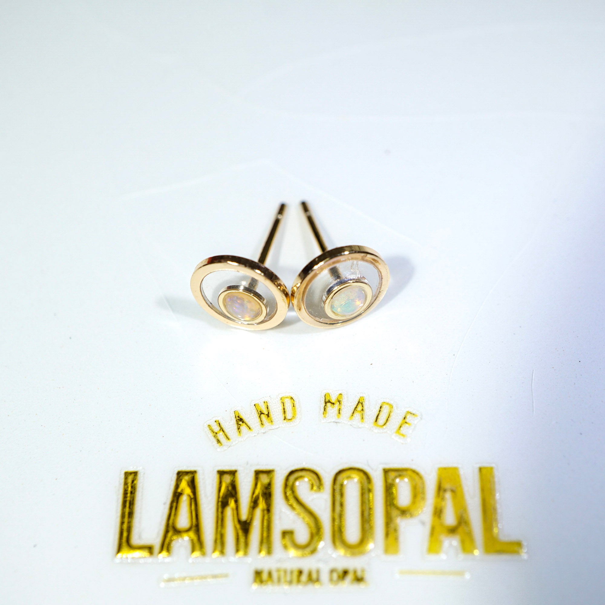 A pair of gold-toned oval stud earrings with natural opal inlays, displayed on a white surface above a gold 'LAMSOPAL' brand logo.