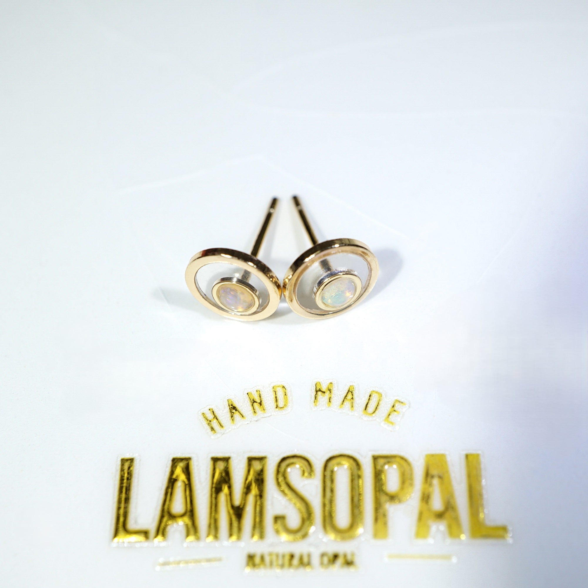A pair of gold-toned stud earrings with circular concentric oval designs and natural opal inlays, displayed on a white surface above a gold 'LAMSOPAL' brand logo.
