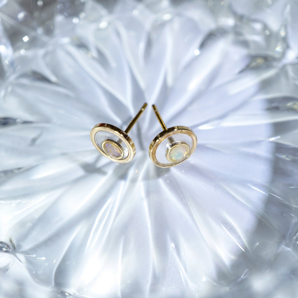 A pair of gold-toned stud earrings with concentric circular cutouts and a small iridescent inlay, displayed on a textured clear glass surface.