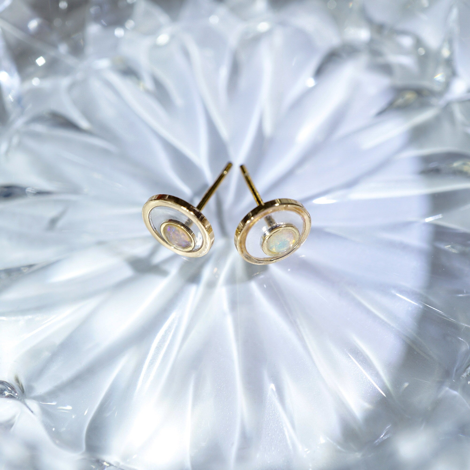 A pair of gold-toned stud earrings with concentric circular cutouts and a small iridescent inlay, displayed on a textured clear glass surface.