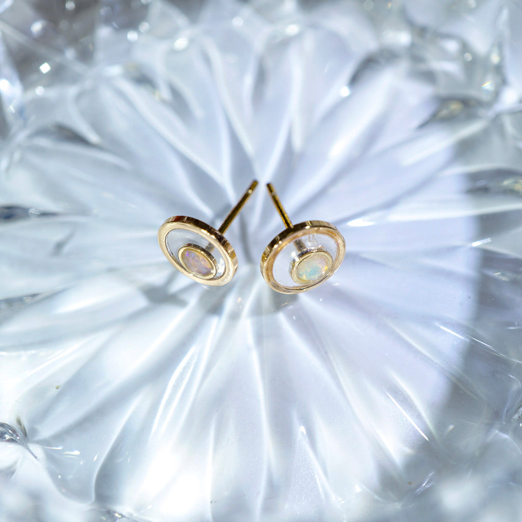 A pair of gold-toned stud earrings with concentric circular cutouts and a small iridescent inlay, displayed on a textured clear glass surface.