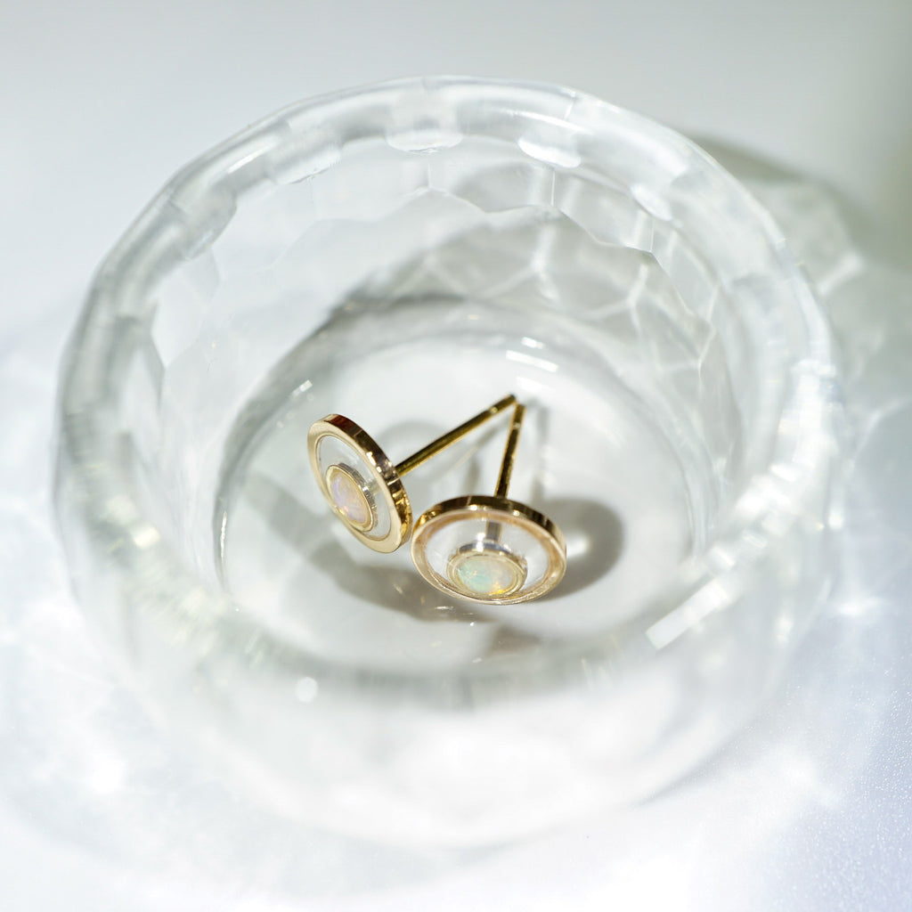 A pair of gold circular stud earrings with iridescent inlay, resting inside a clear faceted glass dish on a white surface.