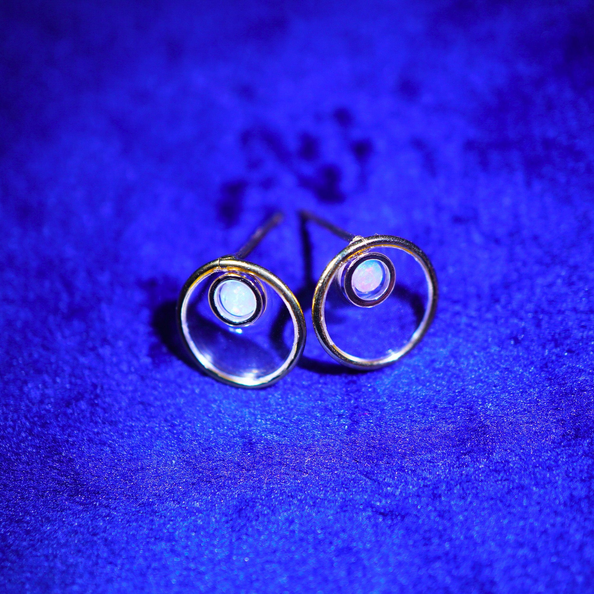 A pair of gold hoop earrings with small oval white inlays, displayed on a textured blue surface.