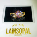 A pair of gold-toned Lamsopal stud earrings displayed on a backing card with a galaxy background, above the brand name 'LAMSOPAL' on a white surface.