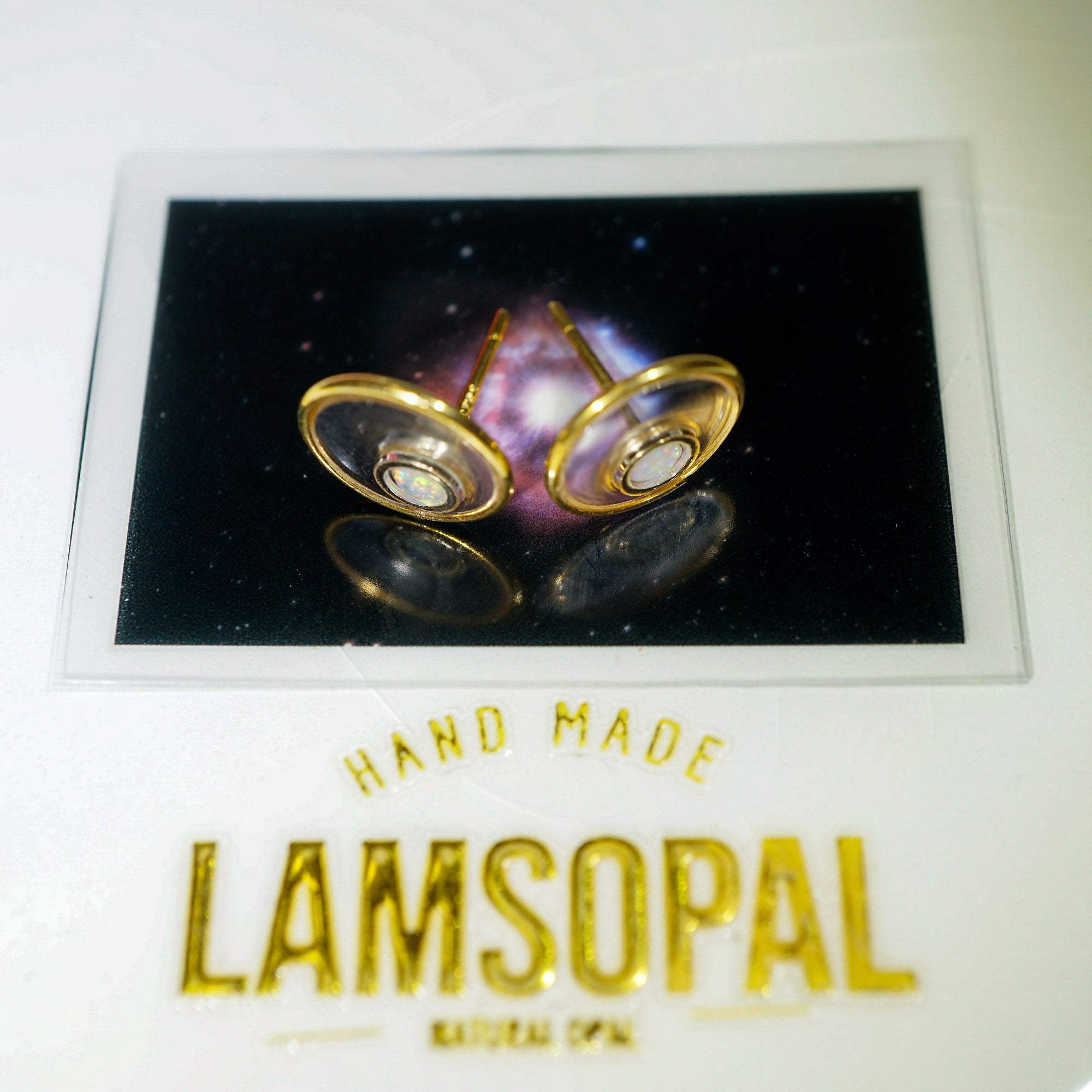 A pair of gold-toned Lamsopal stud earrings displayed on a backing card with a galaxy background, above the brand name 'LAMSOPAL' on a white surface.