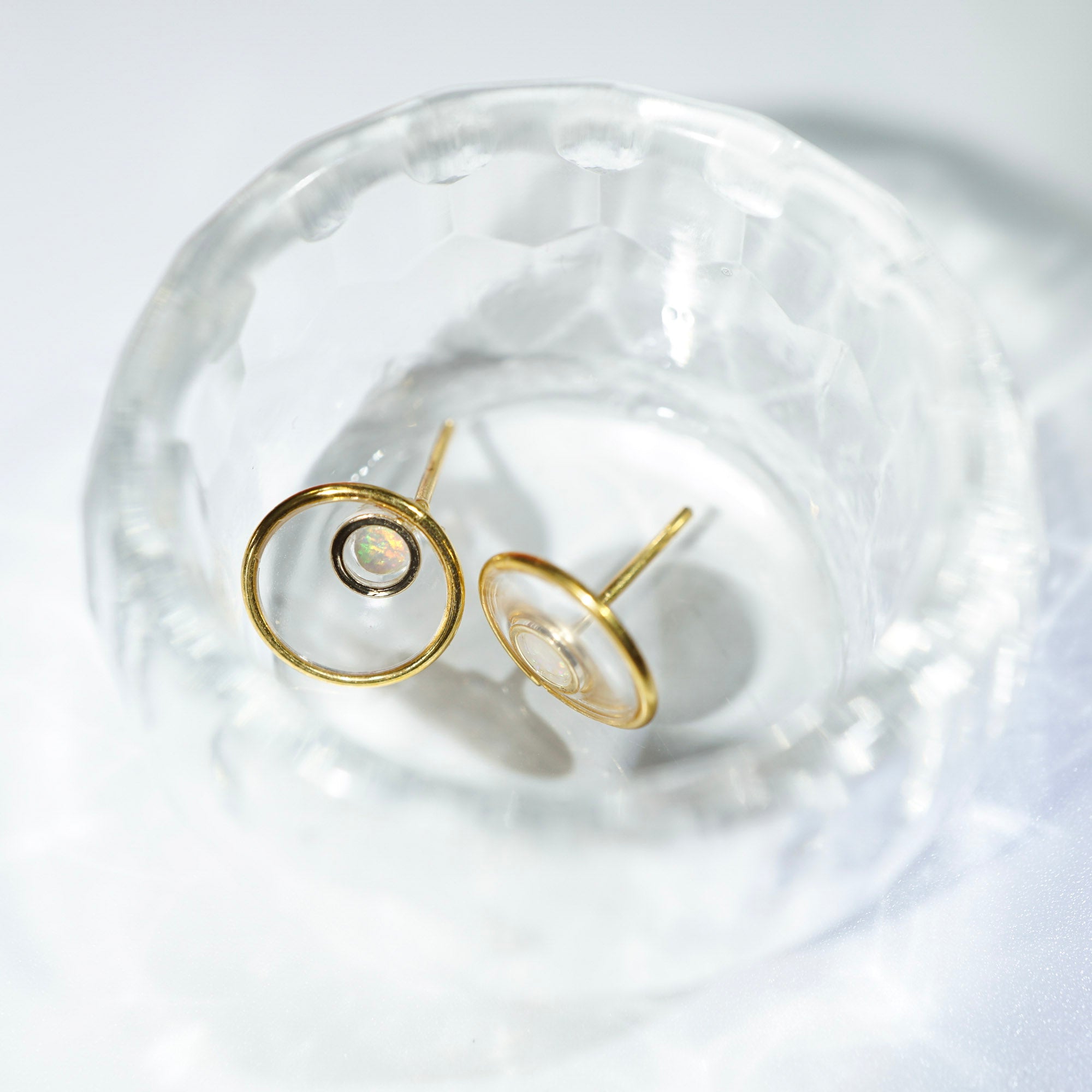 A pair of gold-toned hoop stud earrings with a small black center accent, displayed inside a clear textured glass bowl on a white surface.