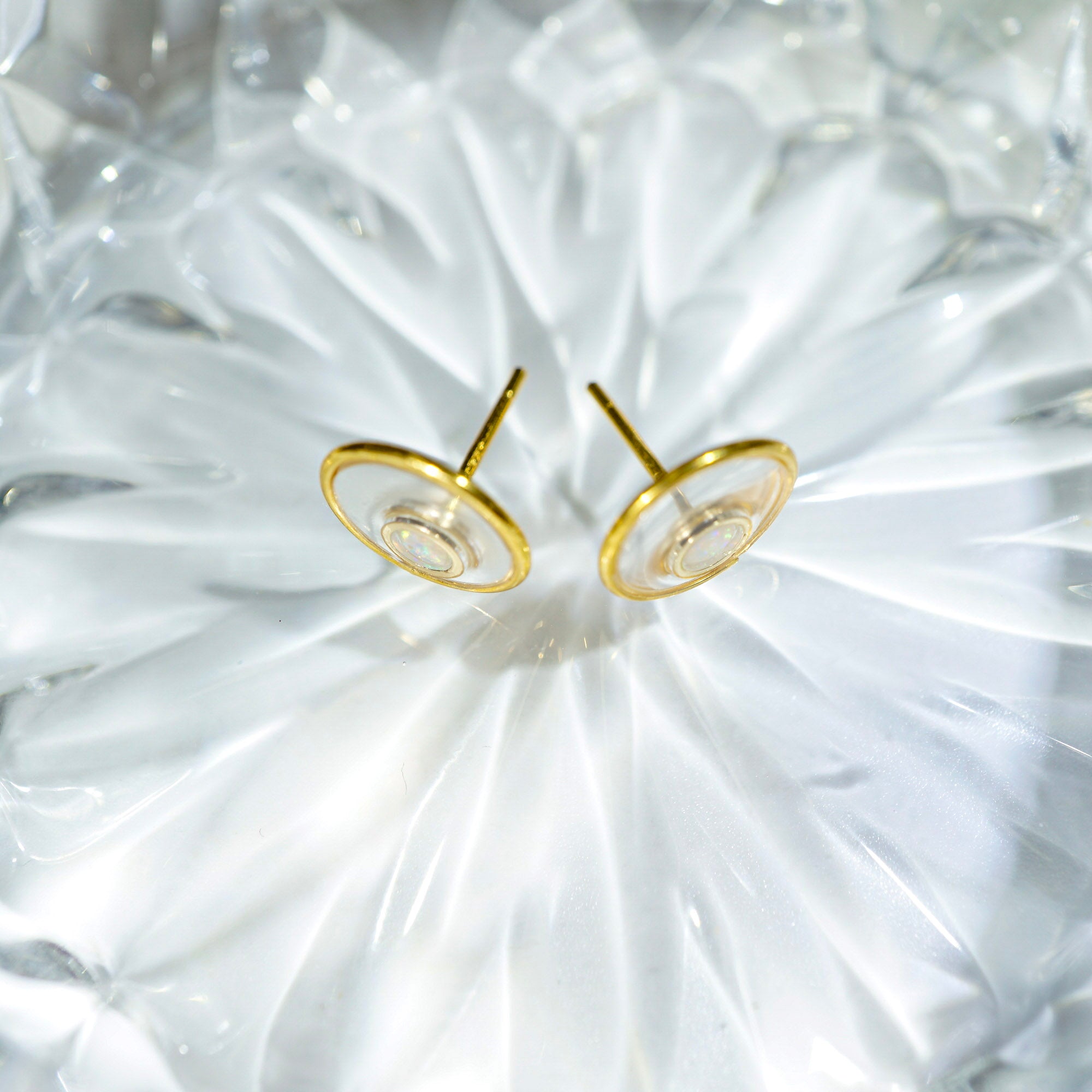 A pair of gold-toned stud earrings with circular disc-shaped fronts, displayed on a textured, translucent white surface.