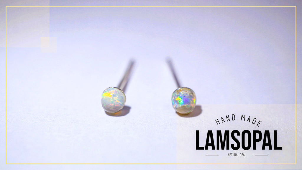 A pair of Lamsopal natural opal stud earrings with silver posts, displayed on a light lavender background with a gold border and a brand logo overlay in the bottom right corner.