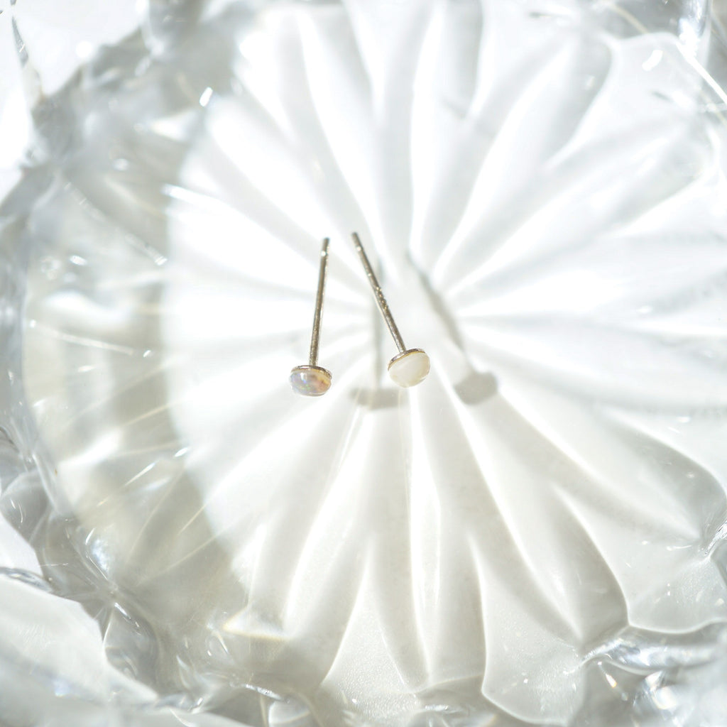 A pair of minimalist stud earrings with small oval pearl-like beads on thin gold-toned posts, resting on a textured clear glass dish.