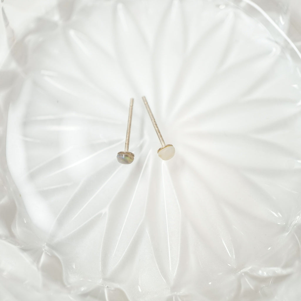 Two small gold stud earrings with round dome-shaped tops, one in a metallic gold finish and one in a champagne gold finish, displayed on a white textured glass plate.