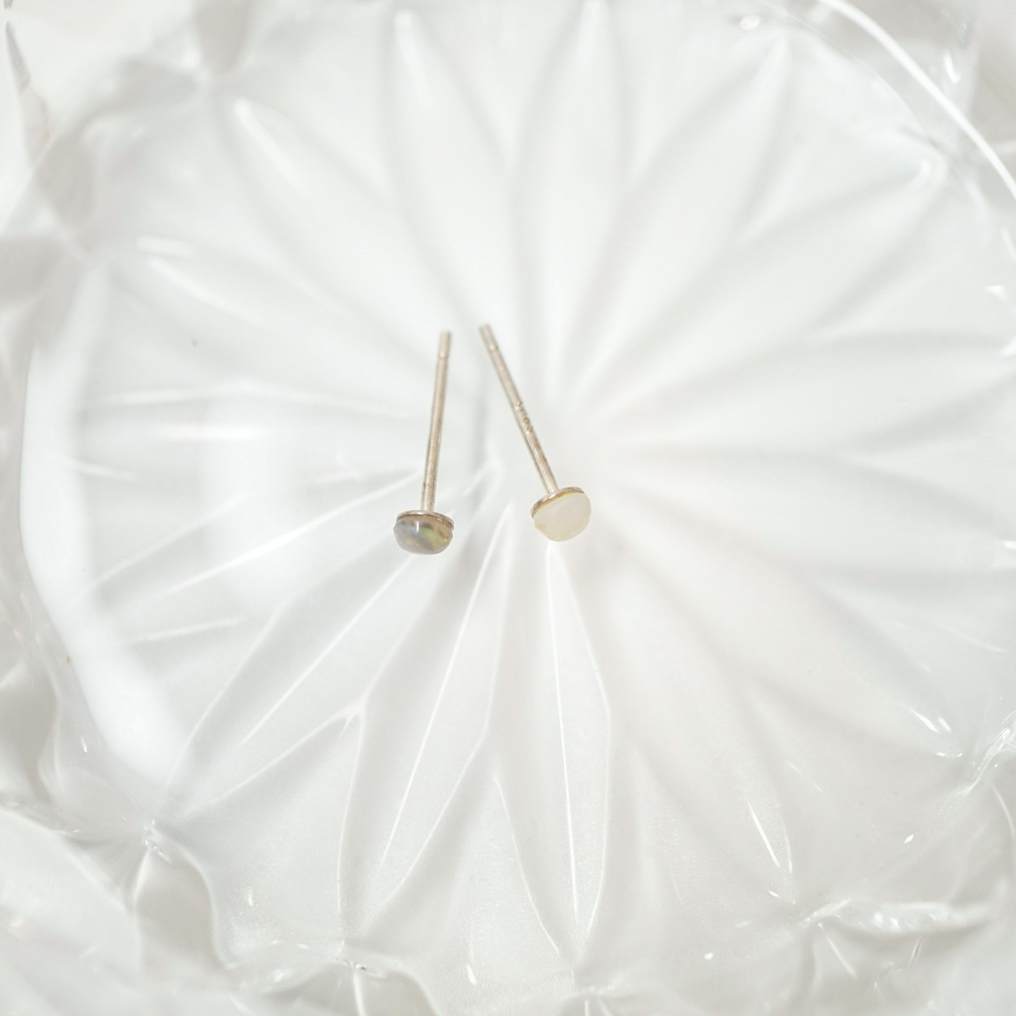 Two small gold stud earrings with round dome-shaped tops, one in a metallic gold finish and one in a champagne gold finish, displayed on a white textured glass plate.