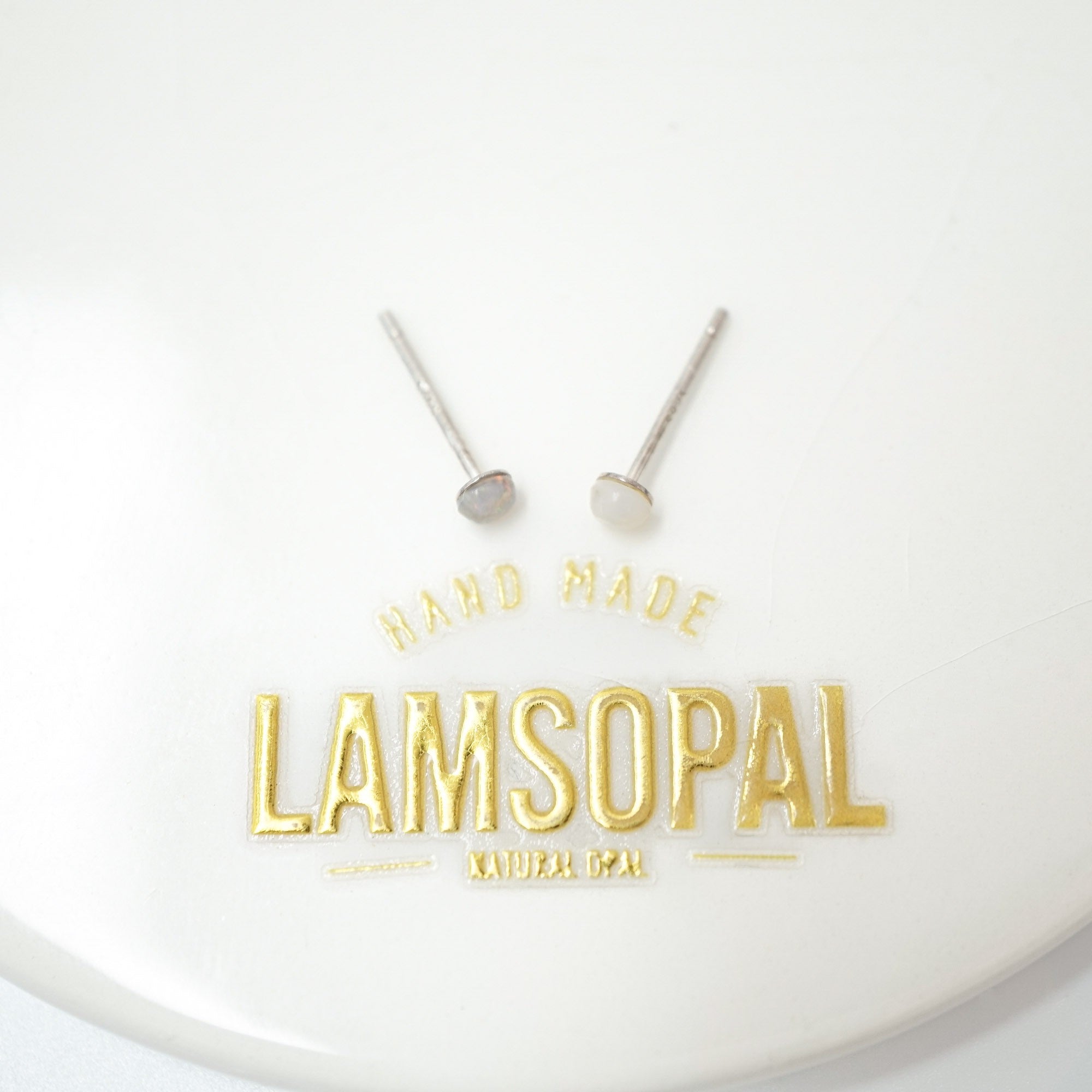 Two silver-toned post earrings with oval clear stones are displayed on a white surface above gold foil branding that reads 'HAND MADE LAMSOPAL NATURAL OPAL.