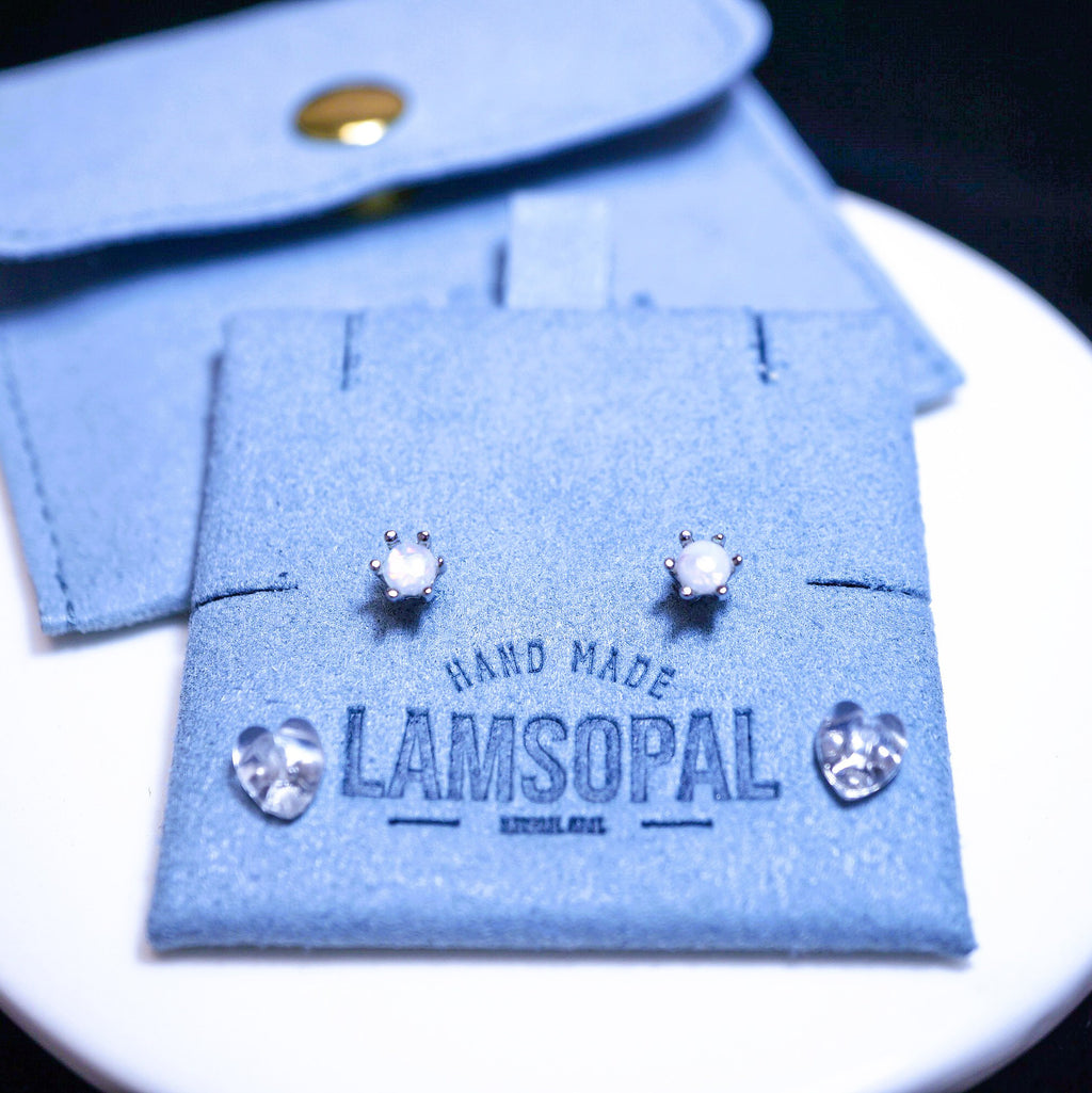 A pair of silver-toned stud earrings with clear stones displayed on a light blue felt jewelry card that reads "HAND MADE LAMSOPAL," with a matching blue felt pouch behind it on a white plate.