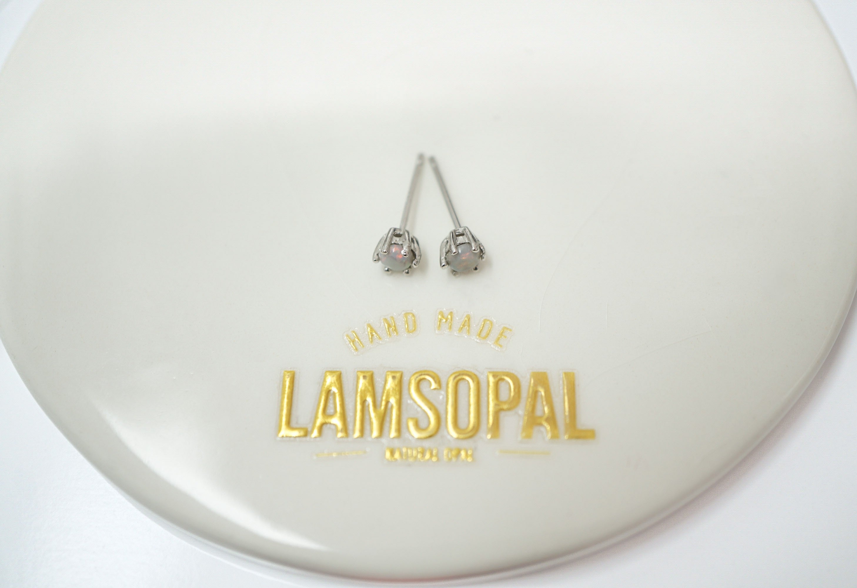 A pair of silver-toned stud earrings with small clear stones and pink accents, displayed on a glossy white oval plate with gold 'LAMSOPAL' branding.