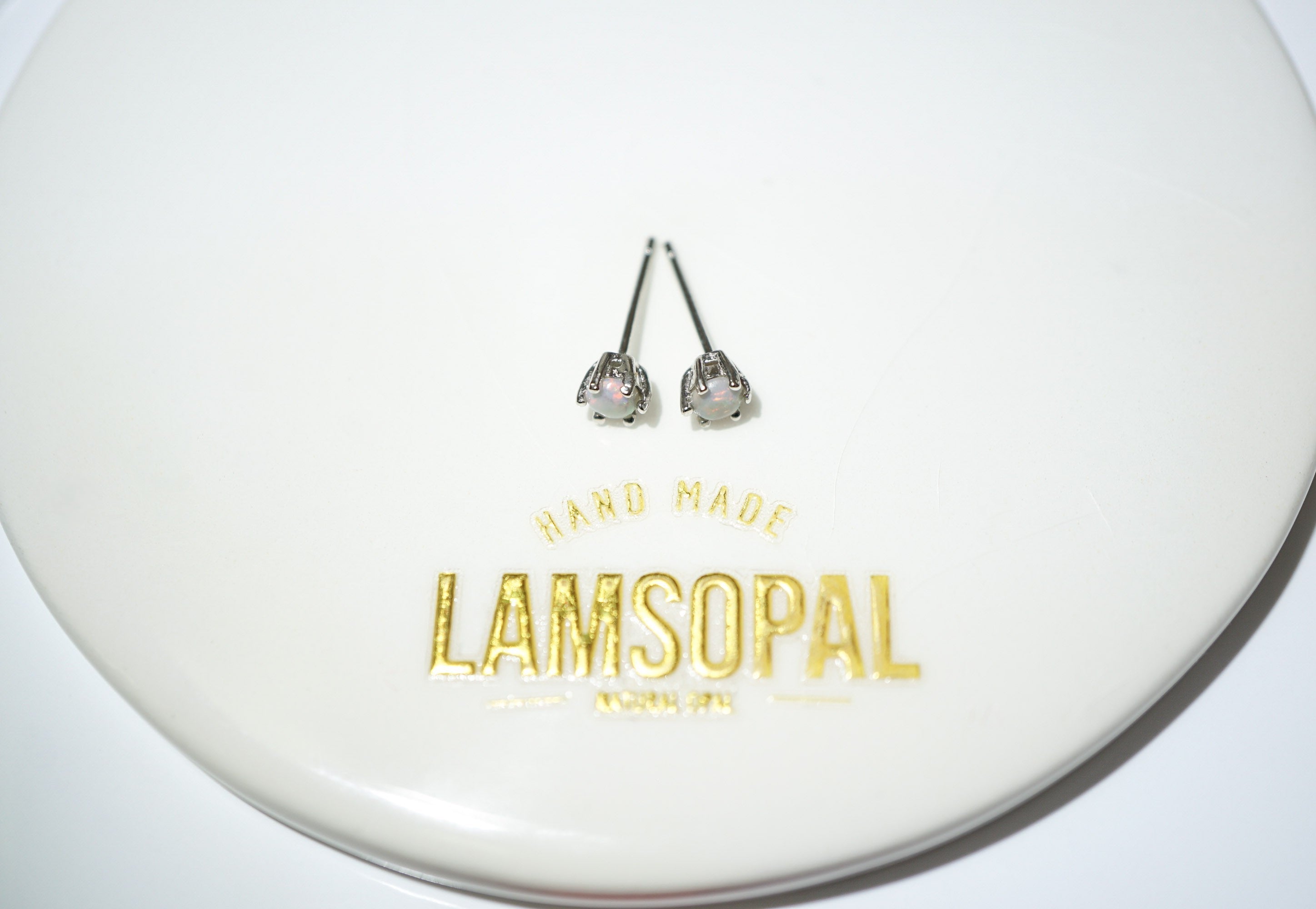 A pair of silver-toned stud earrings with clear faceted gemstones, displayed on a white circular plate printed with the brand name 'LAMSOPAL'.