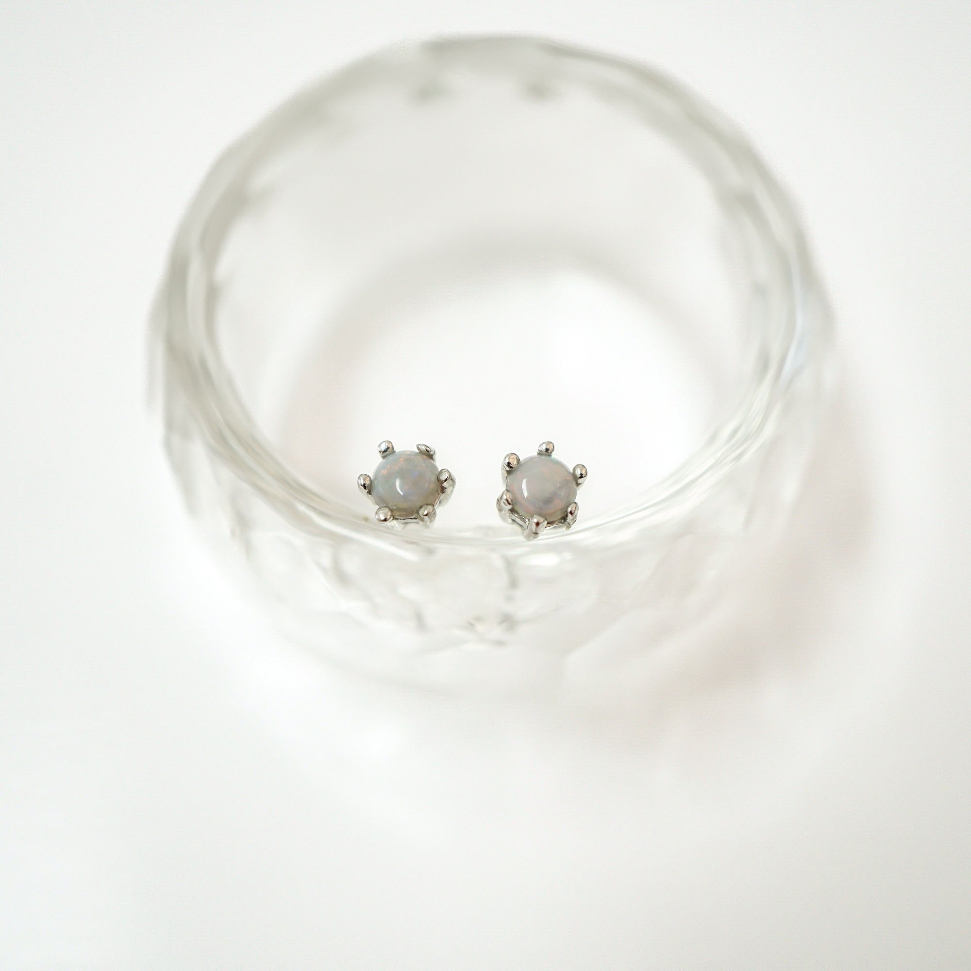 A pair of silver-toned stud earrings with oval milky white stones, displayed inside a clear textured glass bowl on a white background.