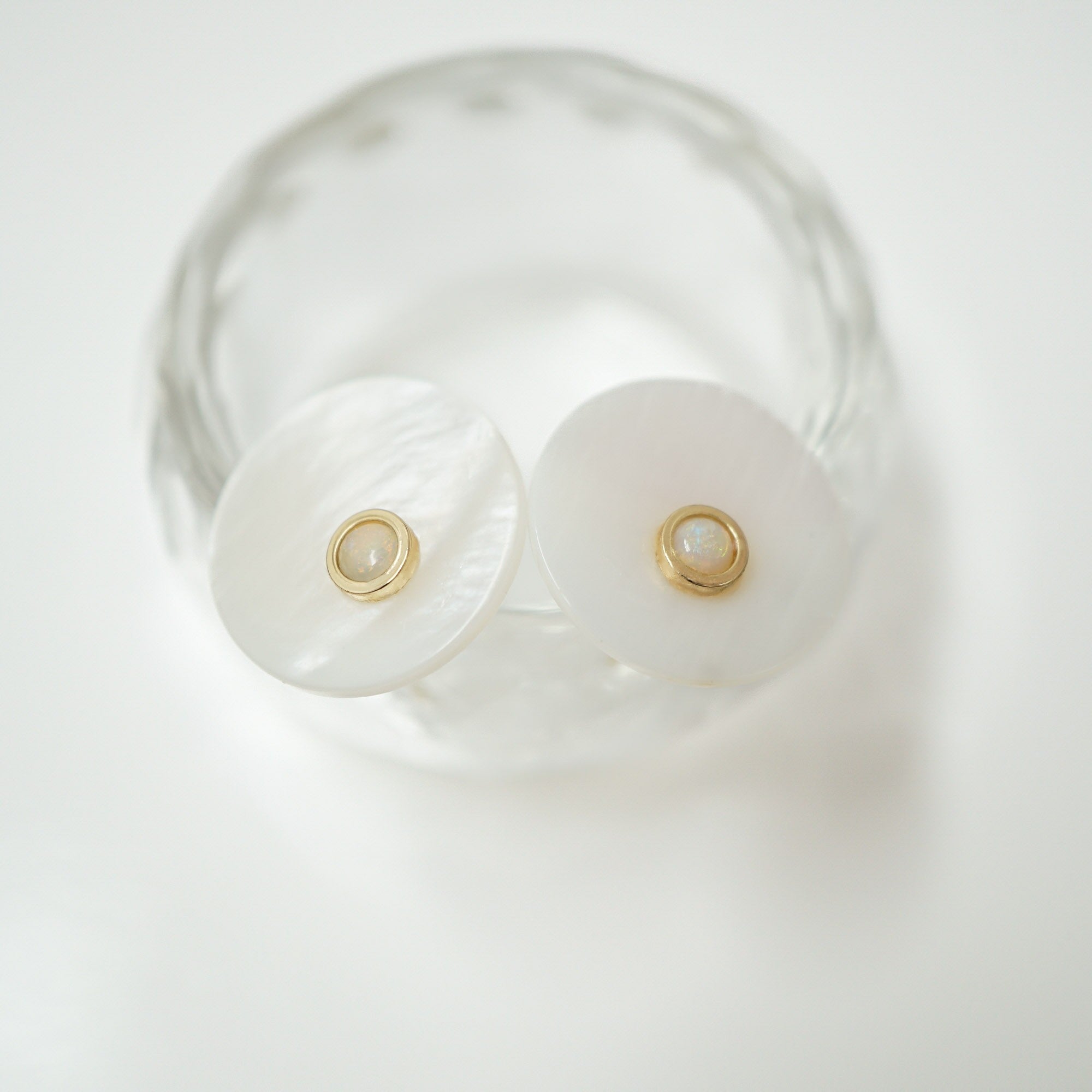 A pair of white disc-shaped stud earrings with a gold bezel setting and a small white pearl in the center, displayed on a clear glass dish against a white background.