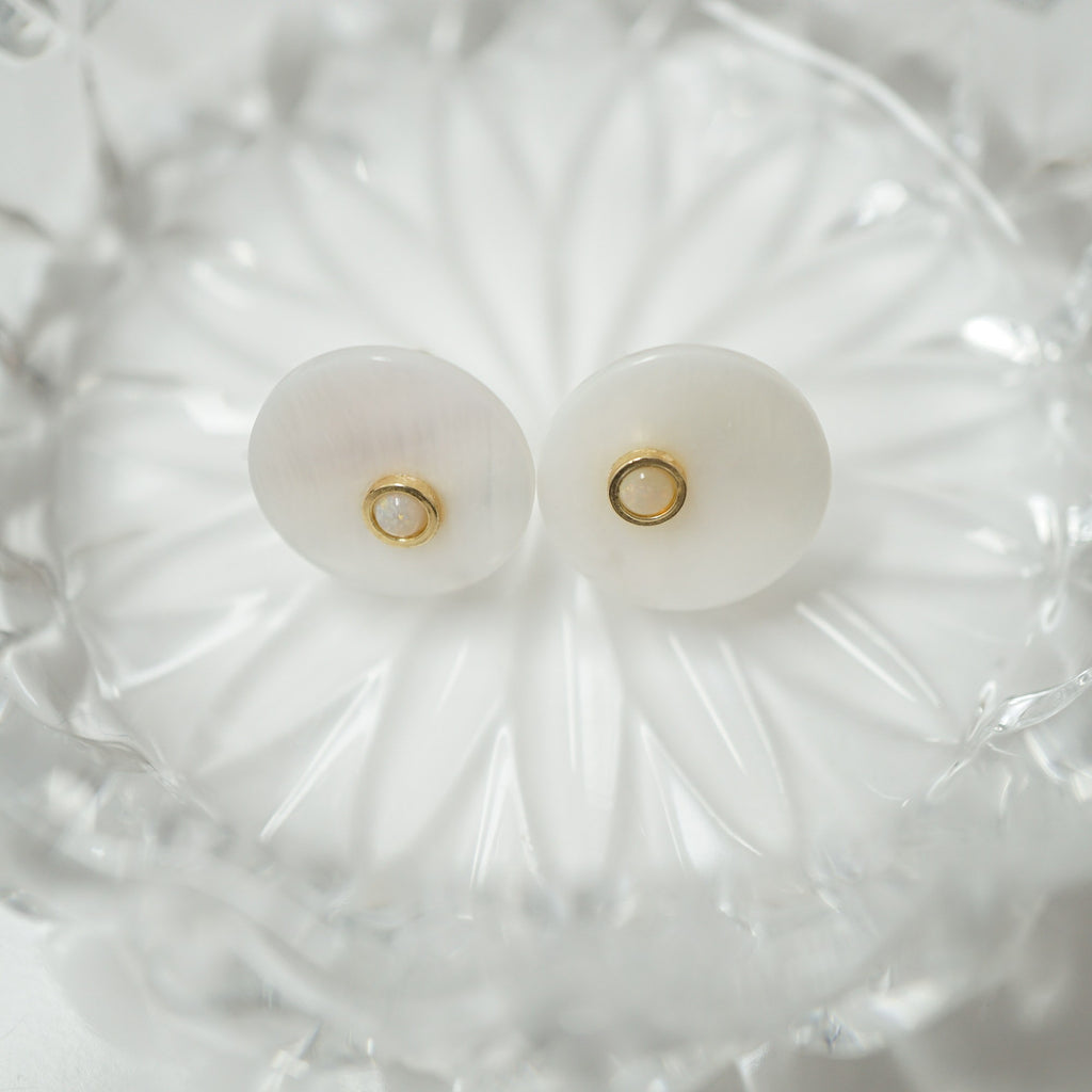 A pair of round white shell stud earrings with small gold center rings, displayed on a clear glass dish.