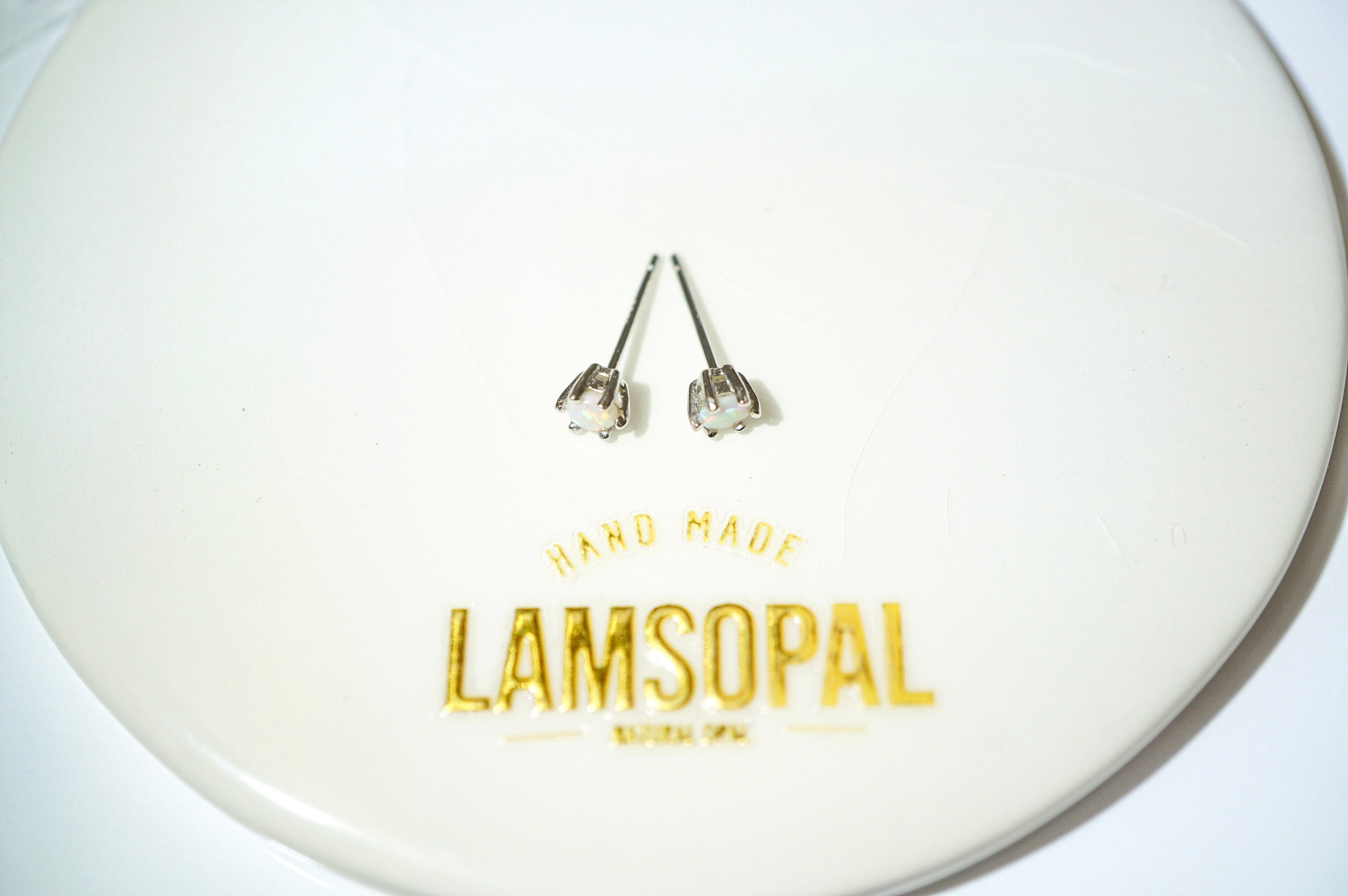 A pair of small silver stud earrings with clear faceted stones, placed on a white circular display plate printed with the brand name 'LAMSOPAL'.