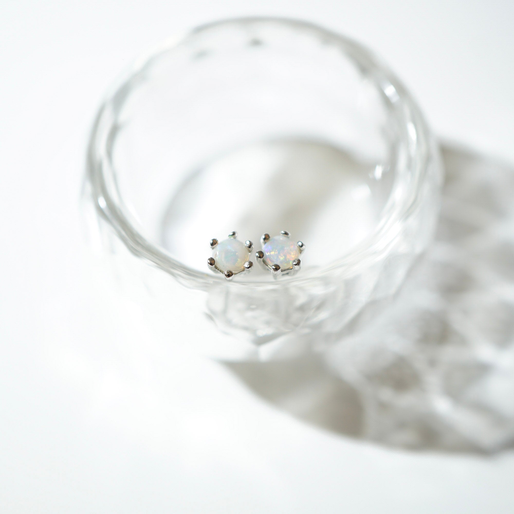 A pair of small silver stud earrings with round iridescent white stones, resting inside a clear textured glass on a white background.