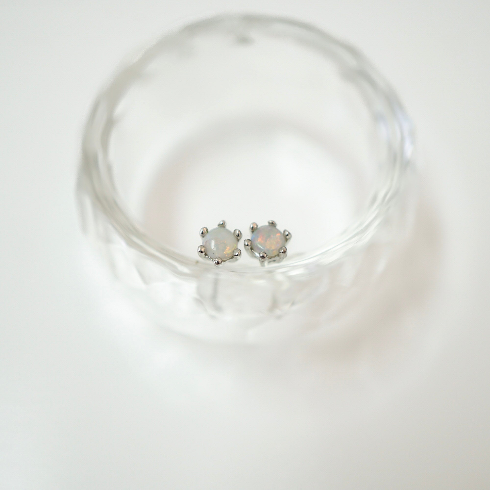 A pair of small silver stud earrings with round iridescent opal-like stones, resting inside a clear textured glass dish on a white background.