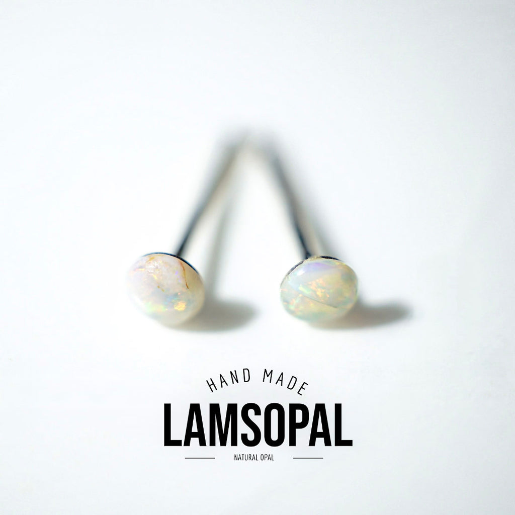 A pair of Lamsopal natural opal stud earrings with round iridescent white opal cabochons on black posts, displayed on a clean white background with a brand logo overlay at the bottom.