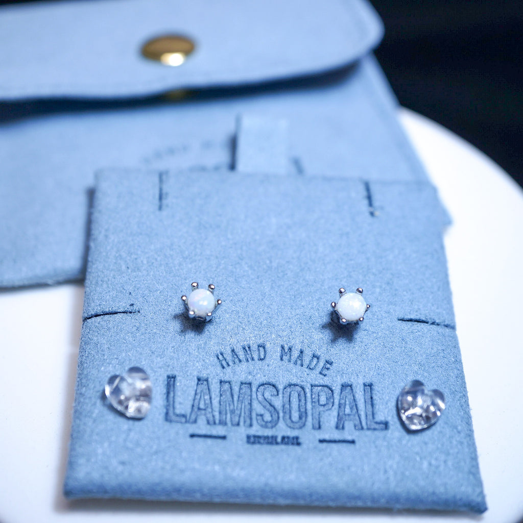 A pair of silver stud earrings with oval white stones displayed on a blue suede jewelry card branded LAMSOPAL, with a matching blue jewelry pouch in the background.