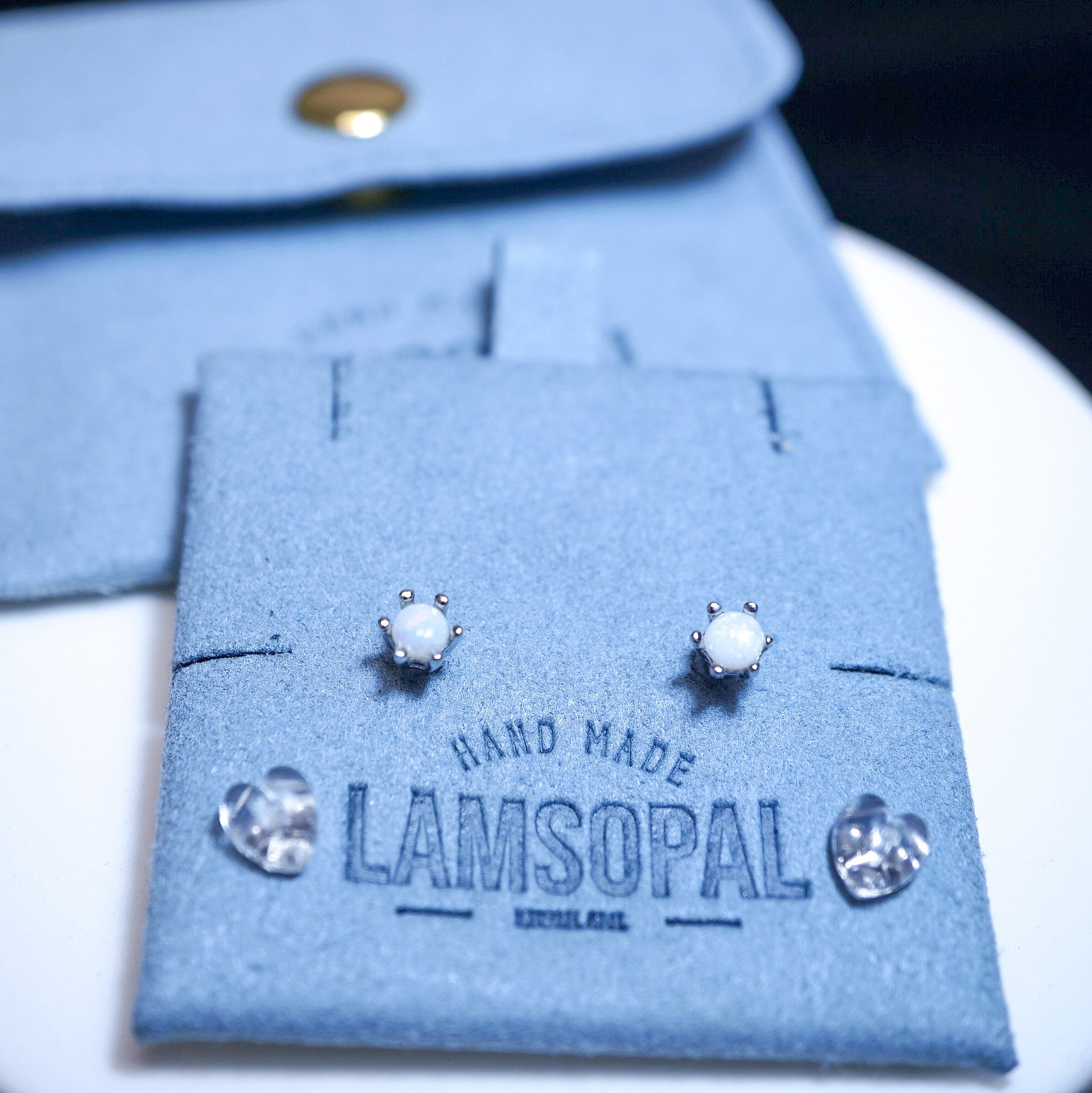 A pair of silver stud earrings with oval white stones displayed on a blue suede jewelry card branded LAMSOPAL, with a matching blue jewelry pouch in the background.