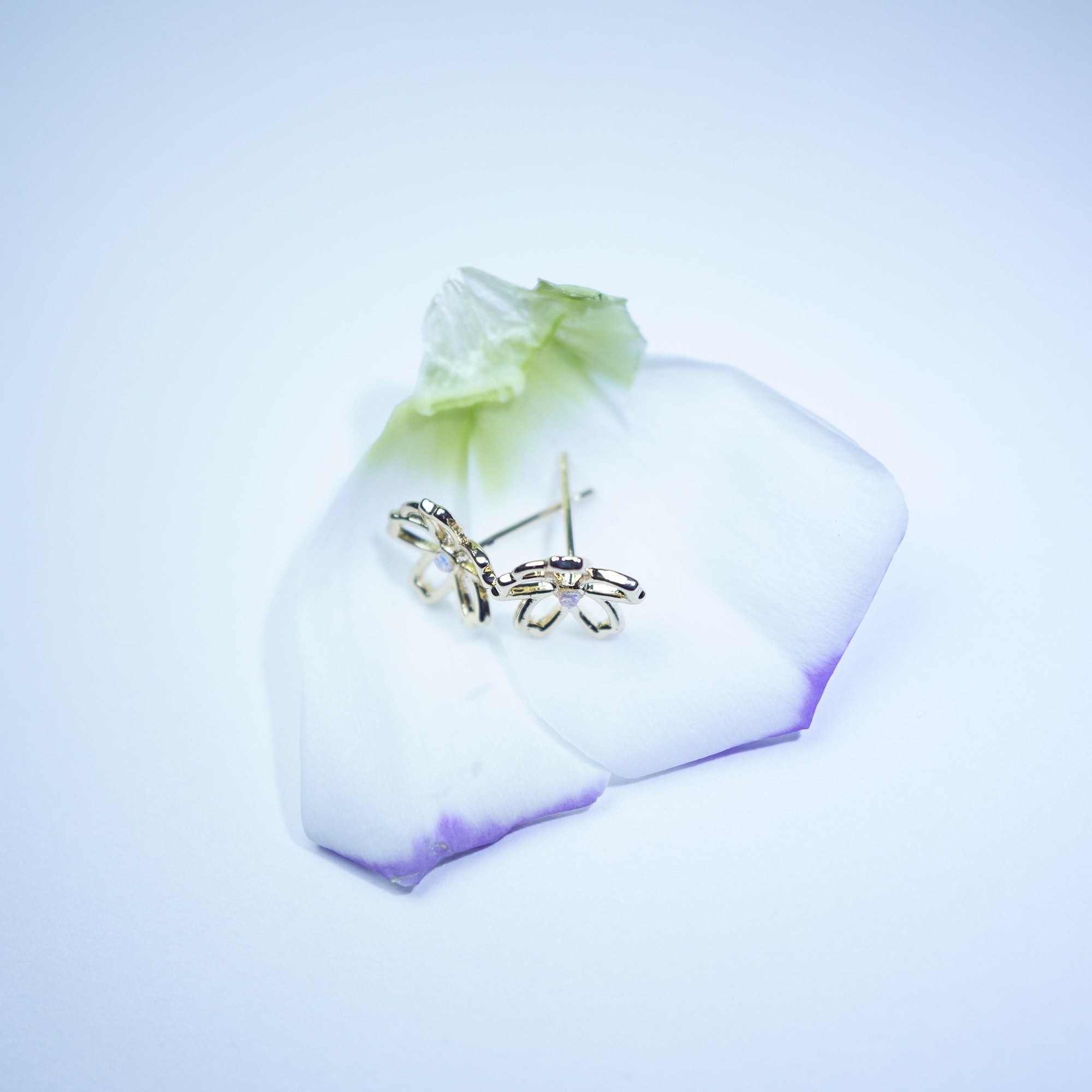 A pair of small gold butterfly stud earrings resting on a white flower petal with a soft blue glow, set against a pale blue background.