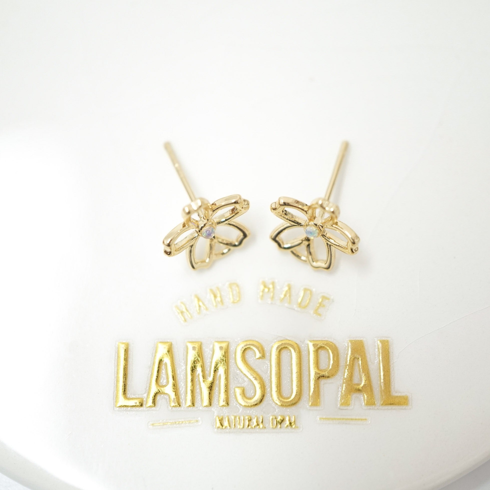 A pair of gold flower-shaped stud earrings with a small clear stone in the center, displayed on a white surface above a gold 'LAMSOPAL' brand logo.