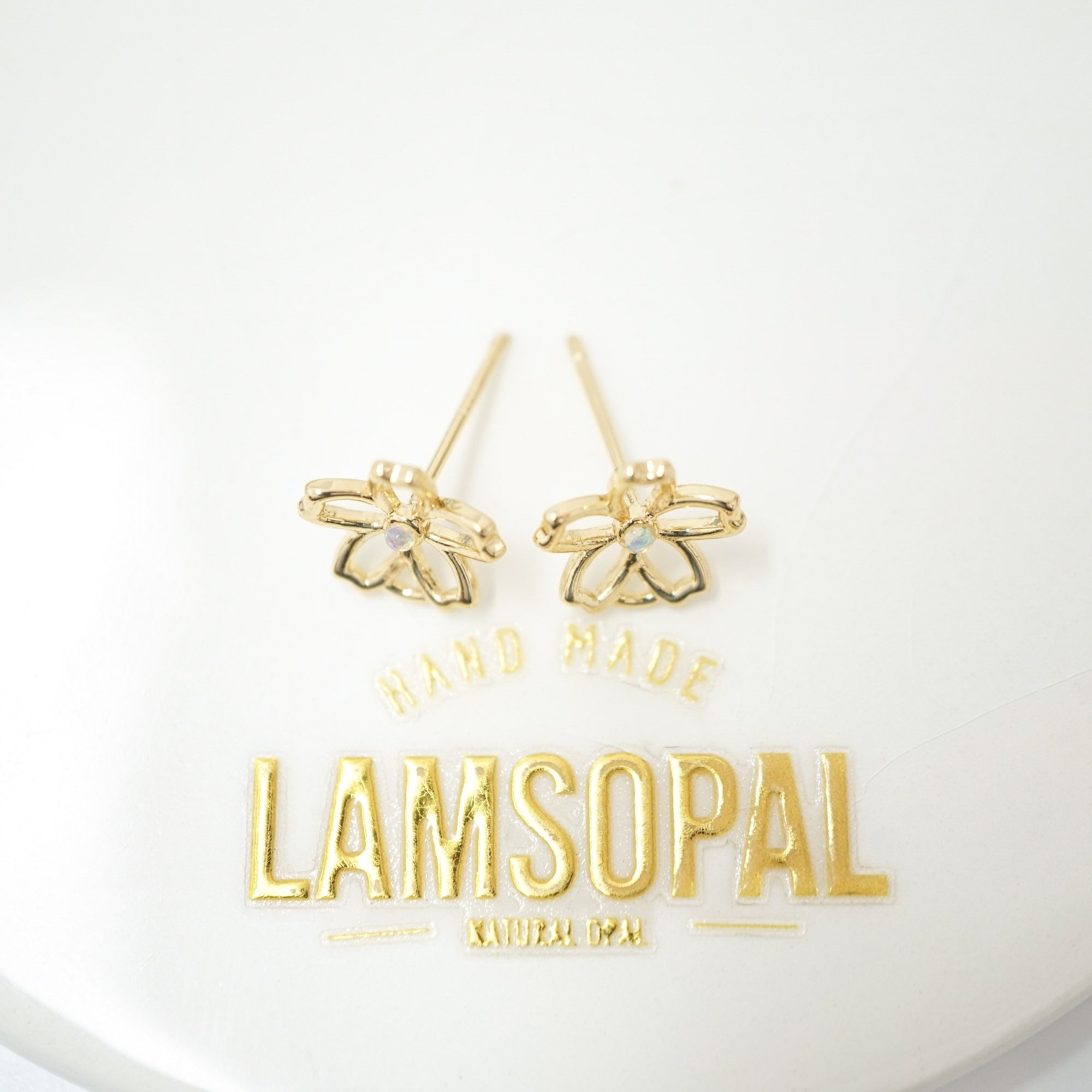 A pair of gold bee stud earrings with a small clear rhinestone in the center, displayed on a white surface above a gold Lamsopal brand plaque.