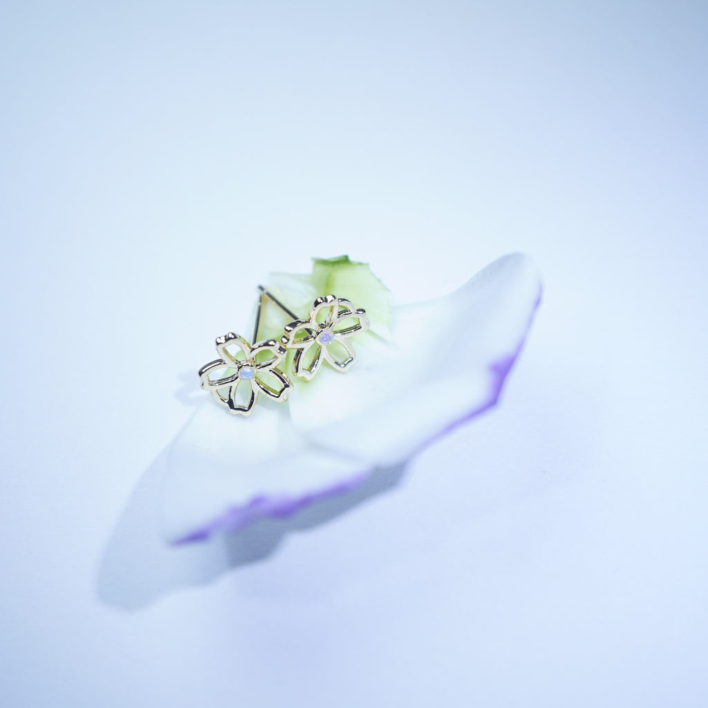 A pair of gold-toned flower-shaped stud earrings resting on a white petal with a soft blue cast against a pale background.