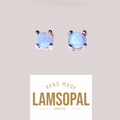 A pair of handmade Lamsopal natural opal stud earrings in silver-toned metal prong settings, shown above a white label with the brand name 'LAMSOPAL'. The opal stones exhibit a milky white color with subtle blue and green iridescent flashes.