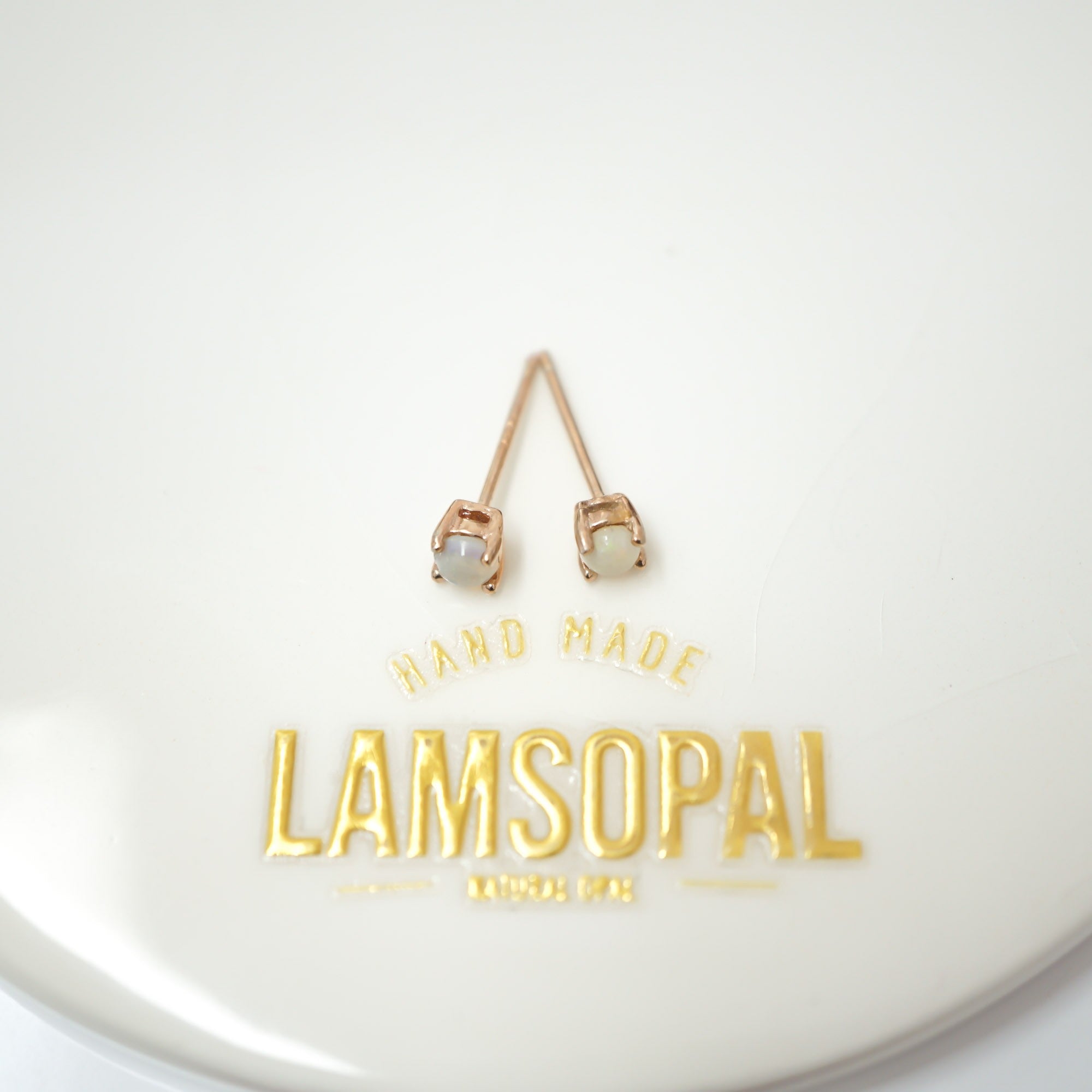 A pair of rose gold stud earrings with oval milky white stones, displayed on a white circular surface with gold foil branding that reads 'HAND MADE LAMSOPAL'.
