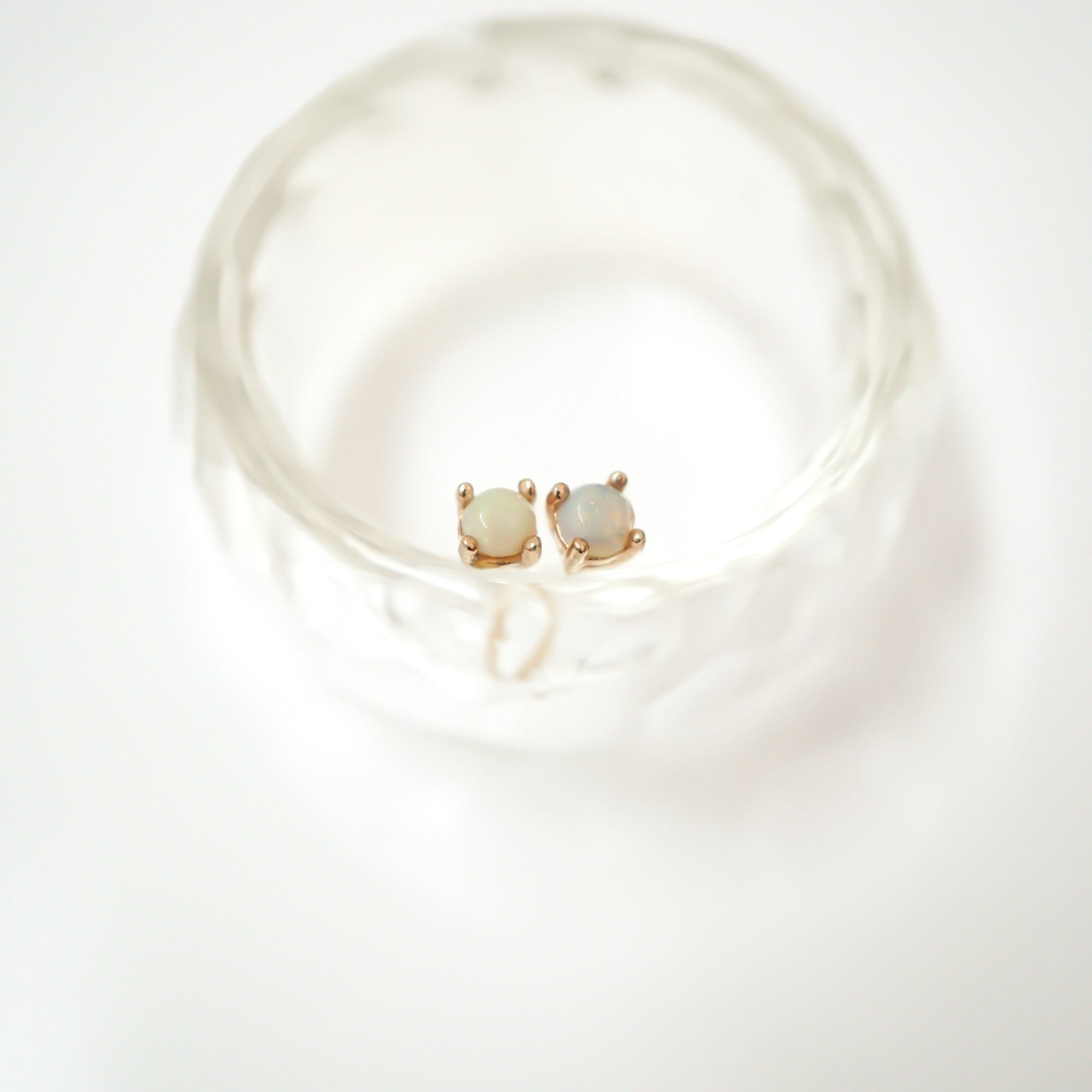 A pair of small gold stud earrings with round pale green opal-like stones, displayed on a clear textured glass dish against a bright white background.
