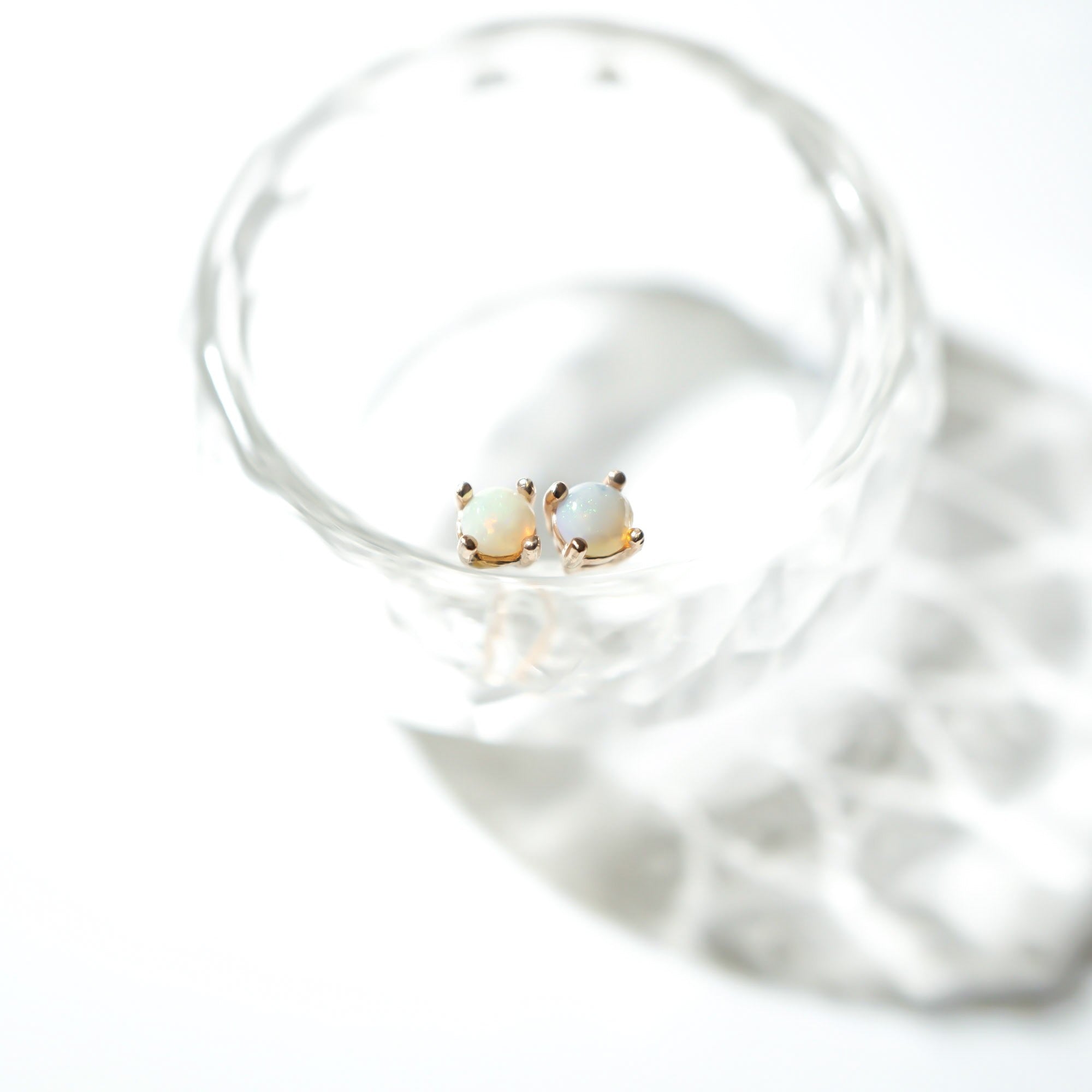 A pair of small gold stud earrings with oval iridescent stones, resting inside a clear faceted glass on a bright white background.