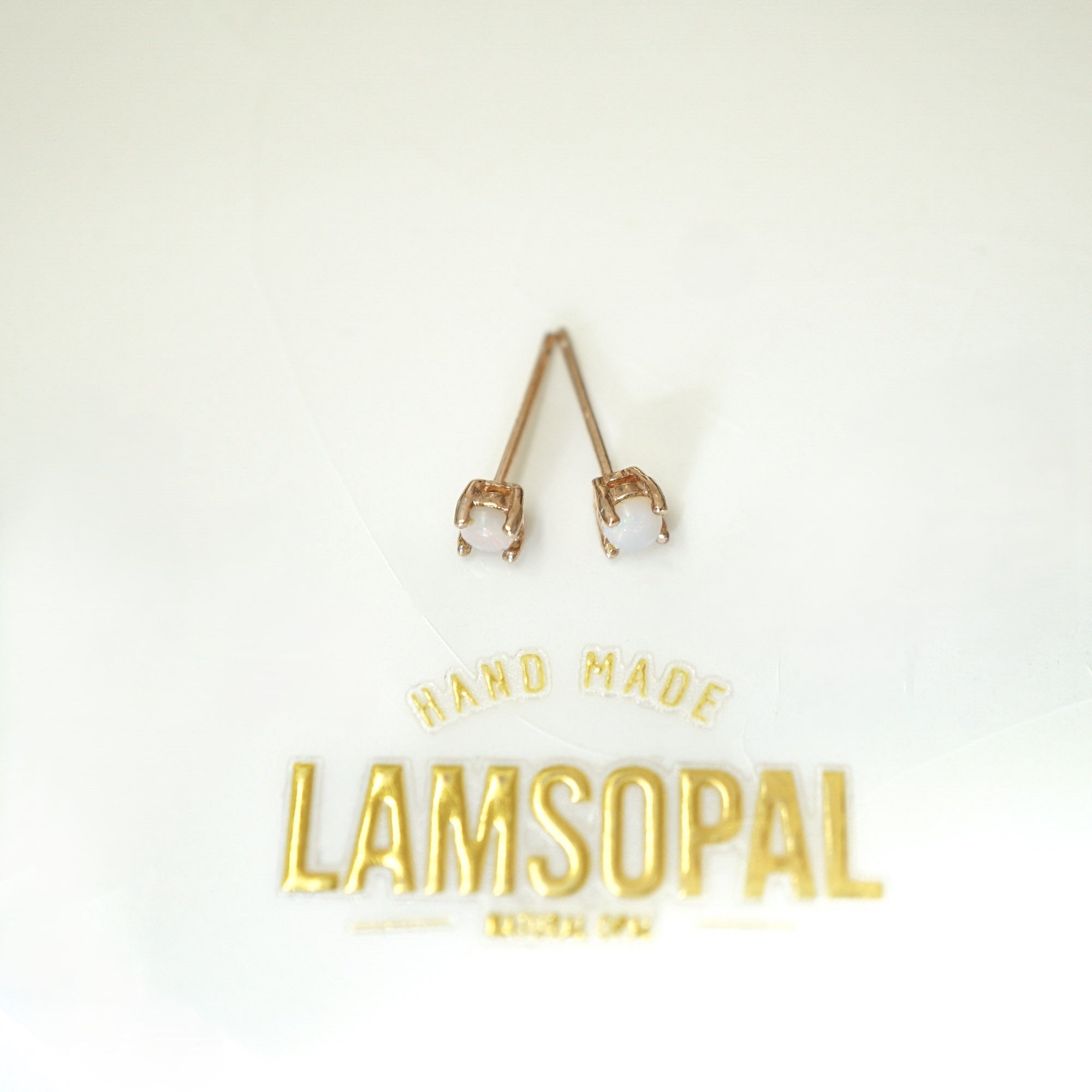 A pair of rose gold stud earrings with clear gemstones in prong settings, displayed on a white background above gold foil branding that reads 'HAND MADE LAMSOPAL'.